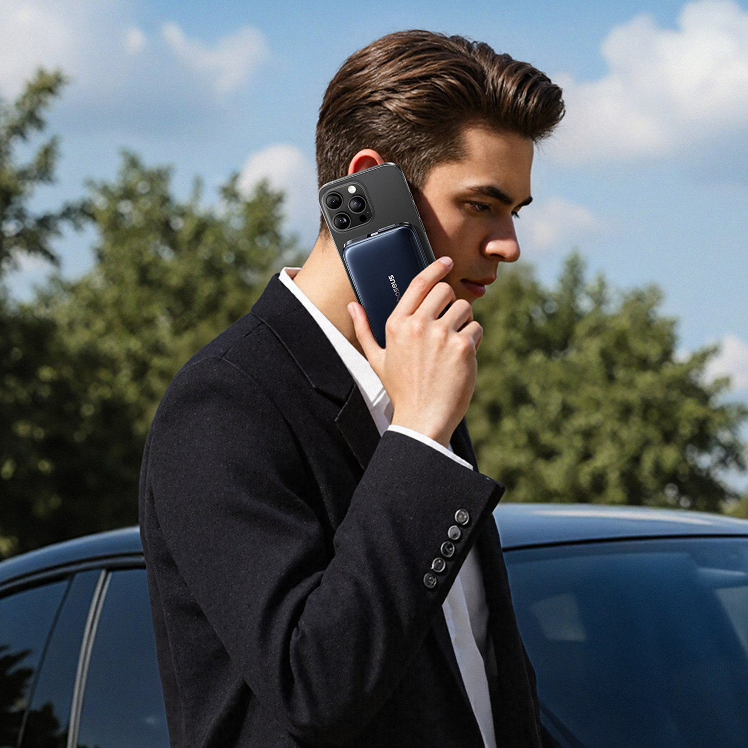 Un hombre en traje sostiene un teléfono con un banco de energía. Está afuera con un coche y árboles al fondo.