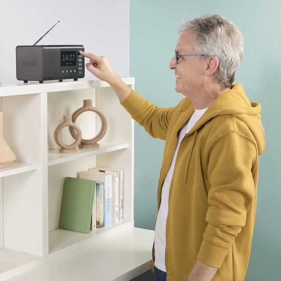 Ein Mann in gelber Jacke bedient ein Radio. Das Radio steht auf einem weißen Regal mit Vasen und Büchern. Im Hintergrund ist eine blaue Wand.