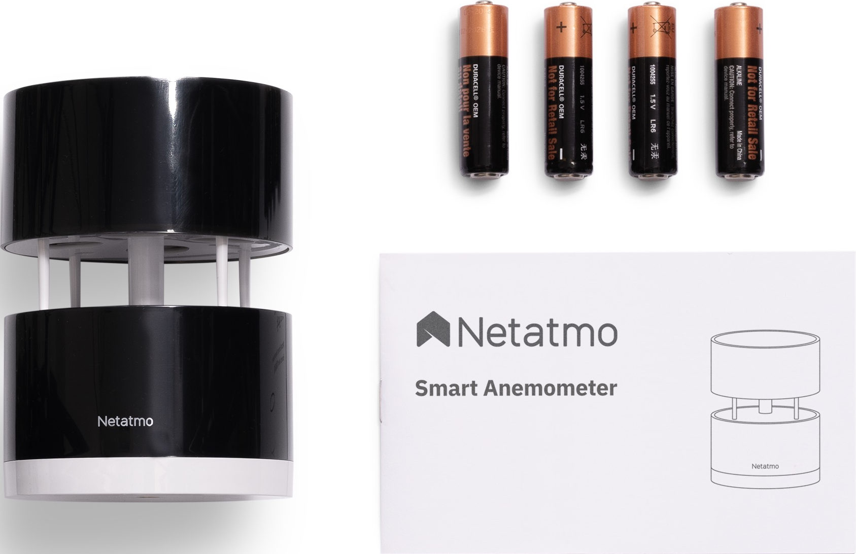L'image présente un anémomètre Netatmo, des piles et un manuel. L'anémomètre est noir et blanc. Les piles sont cuivrées. Le manuel montre un schéma.