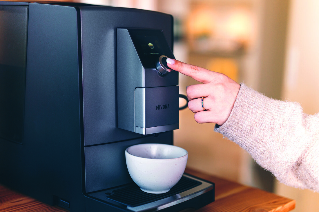 Osoba naciska przycisk na ekspresie do kawy Nivona. Pod wylewką stoi biały kubek. Maszyna stoi na drewnie.