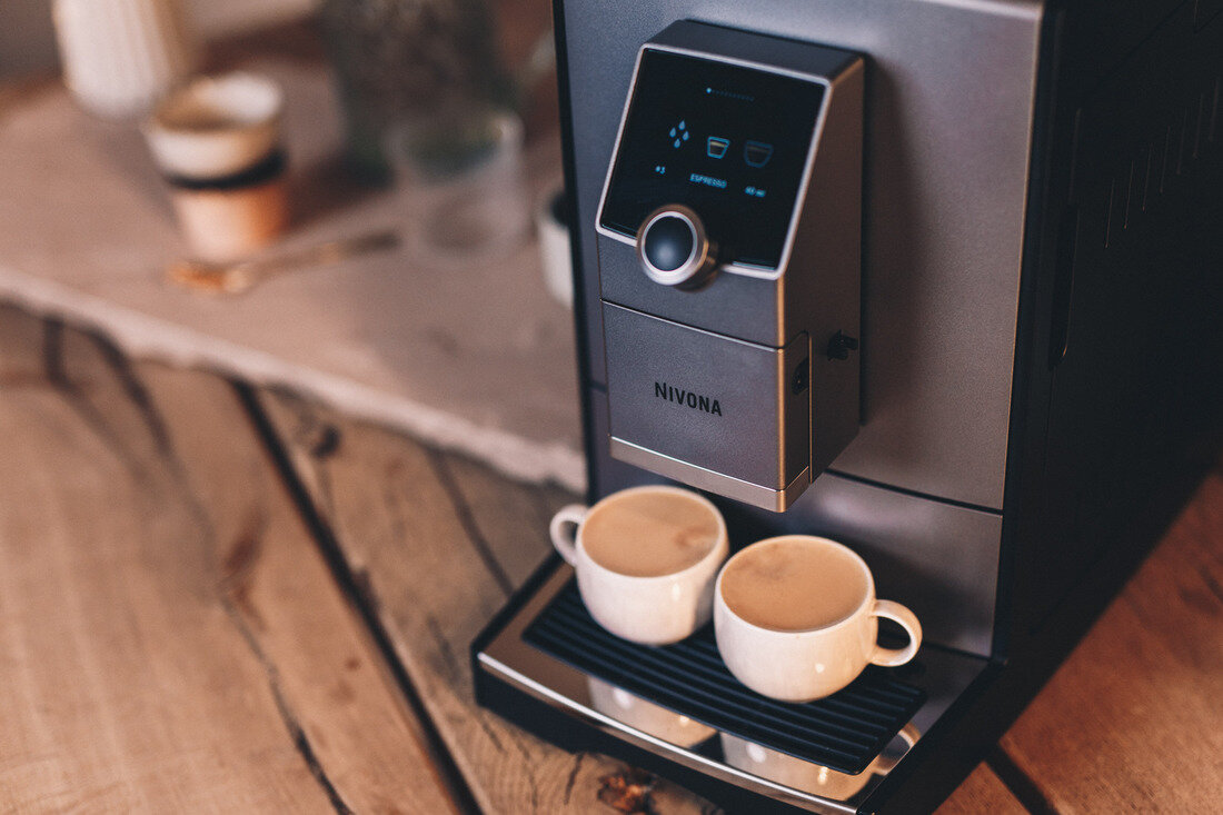 Ekspres do kawy z dwoma filiżankami kawy. Maszyna jest szara i czarna. Drewniany stół i inne przedmioty w tle.