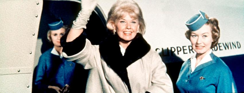 Doris Day winkt mit zwei Stewardessen. Sie trägt einen Mantel mit Pelzkragen. Der Hintergrund hat ein Clipper-Zeichen.