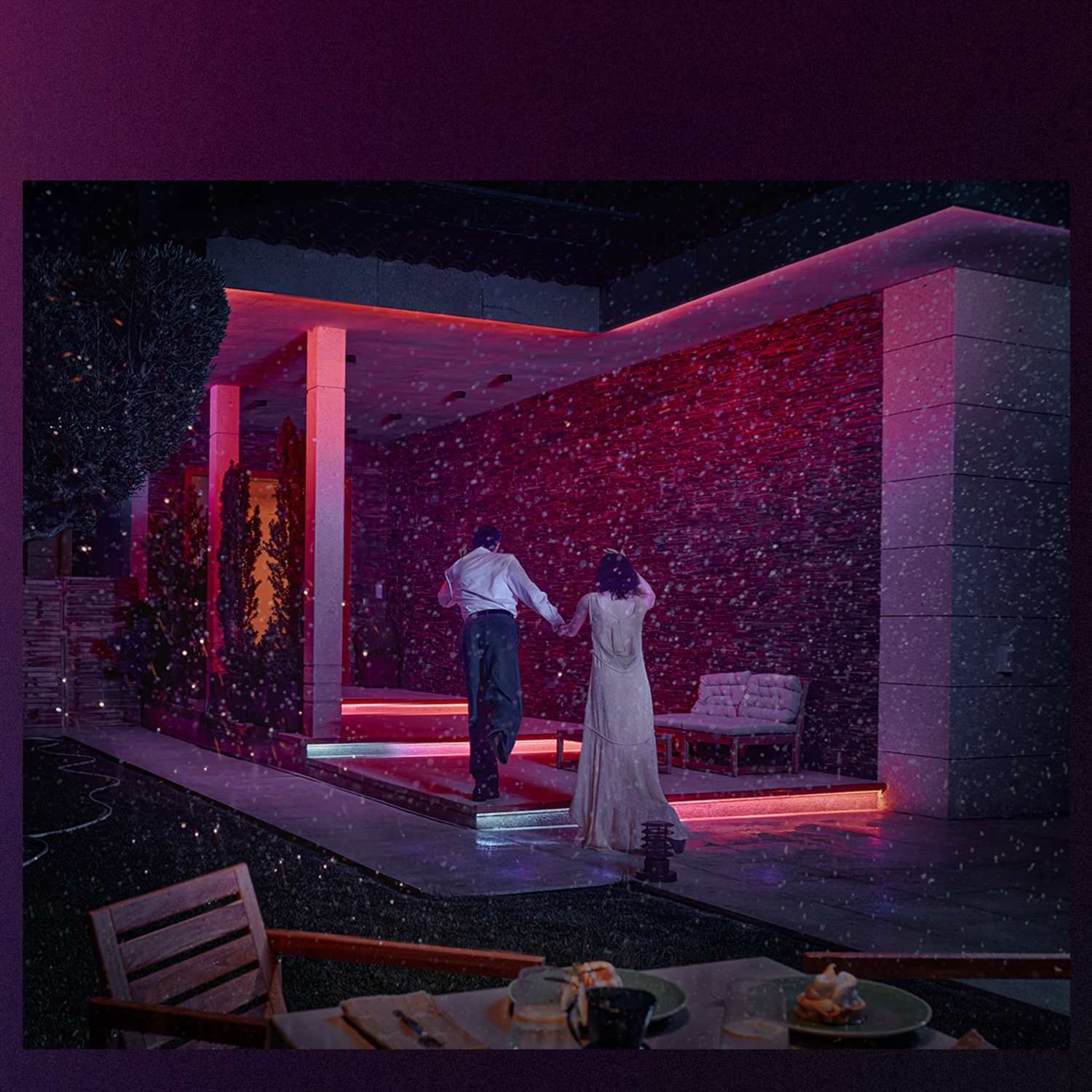 A couple runs toward a lit building with a red wall. Snow falls. A table with food sits in the foreground. 