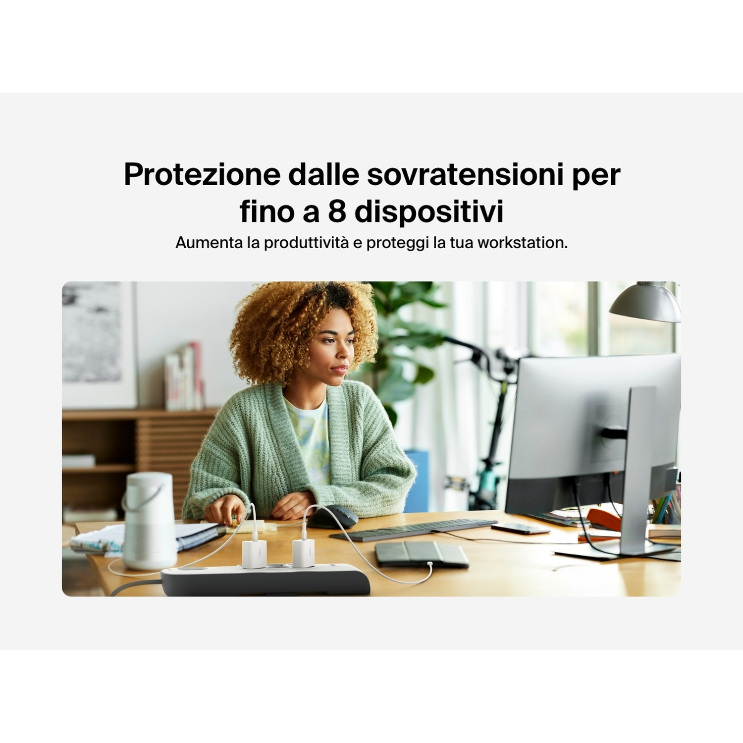 Una donna è seduta a una scrivania, guarda un computer. Nel primo piano c'è una ciabatta con caricabatterie.