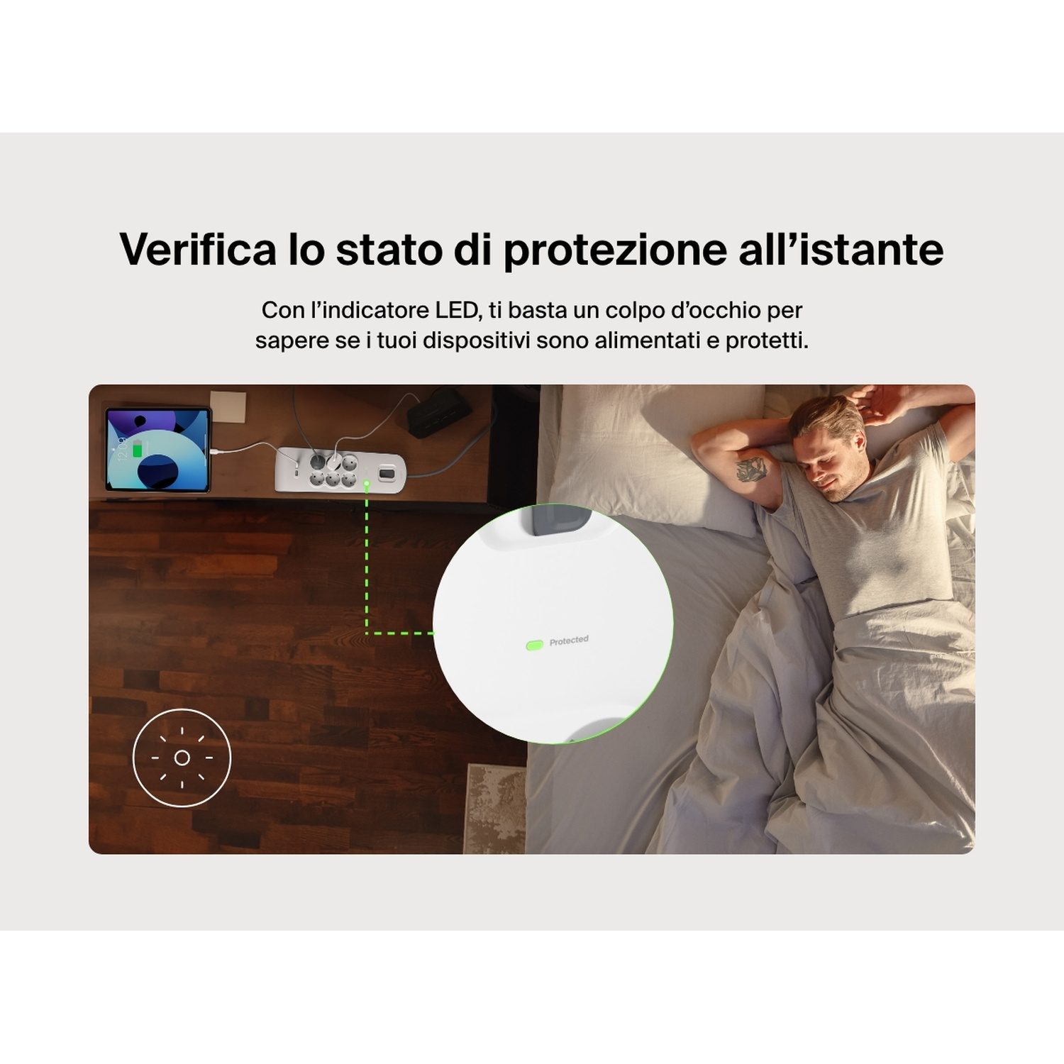 Un uomo a letto, una presa multipla su un tavolo e un tablet sono visibili. La presa multipla ha una luce verde che indica protezione.