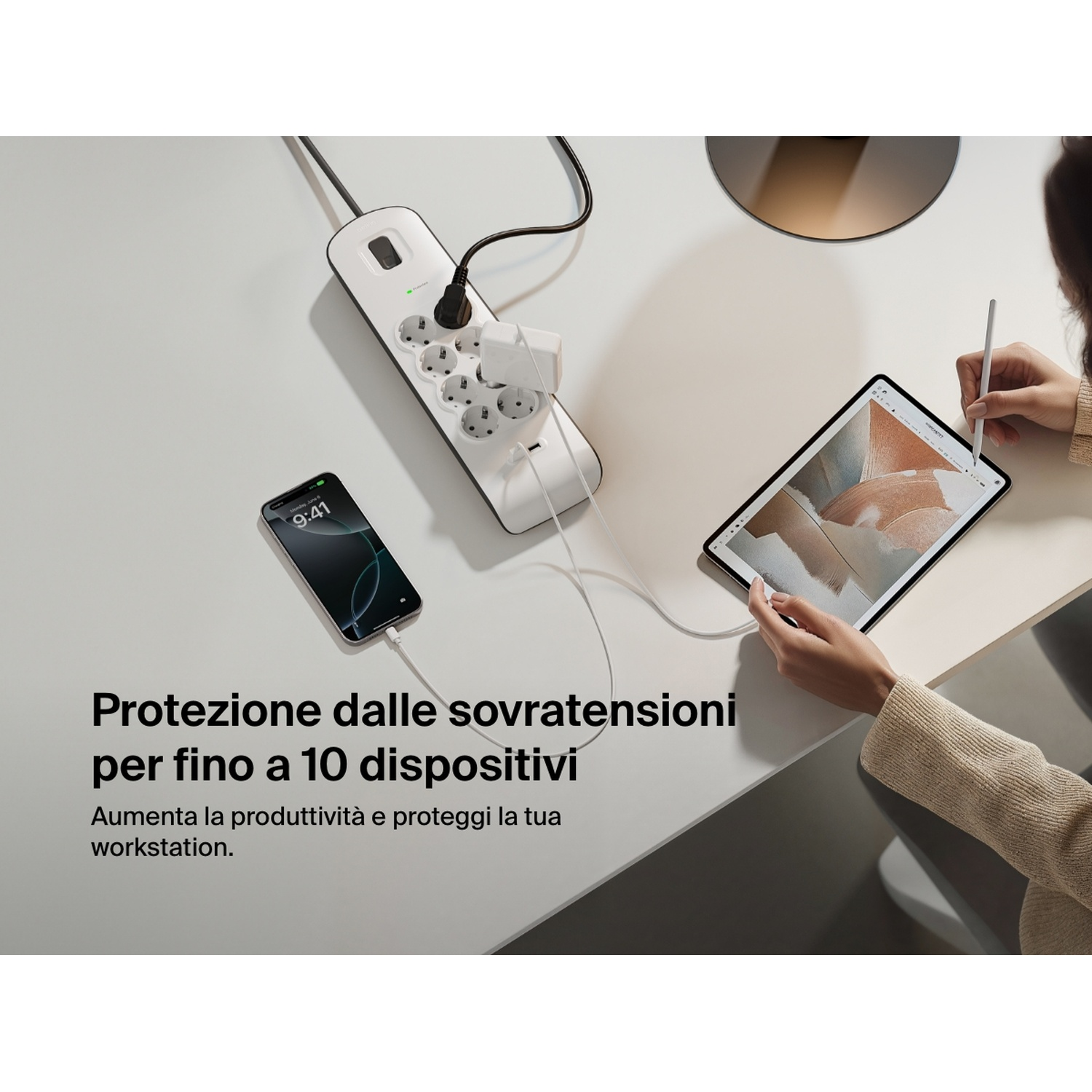Una ciabatta bianca con dispositivi collegati. Un telefono e un tablet sono sulla scrivania con una persona che usa il tablet.