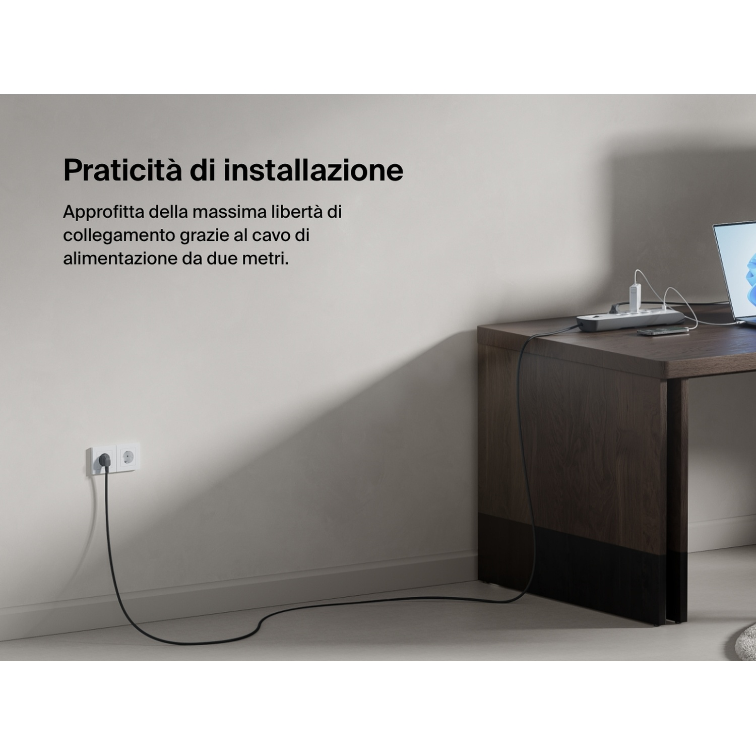 Una presa multipla collegata a una presa a muro. Un laptop è su una scrivania di legno. Testo: Praticità di installazione.