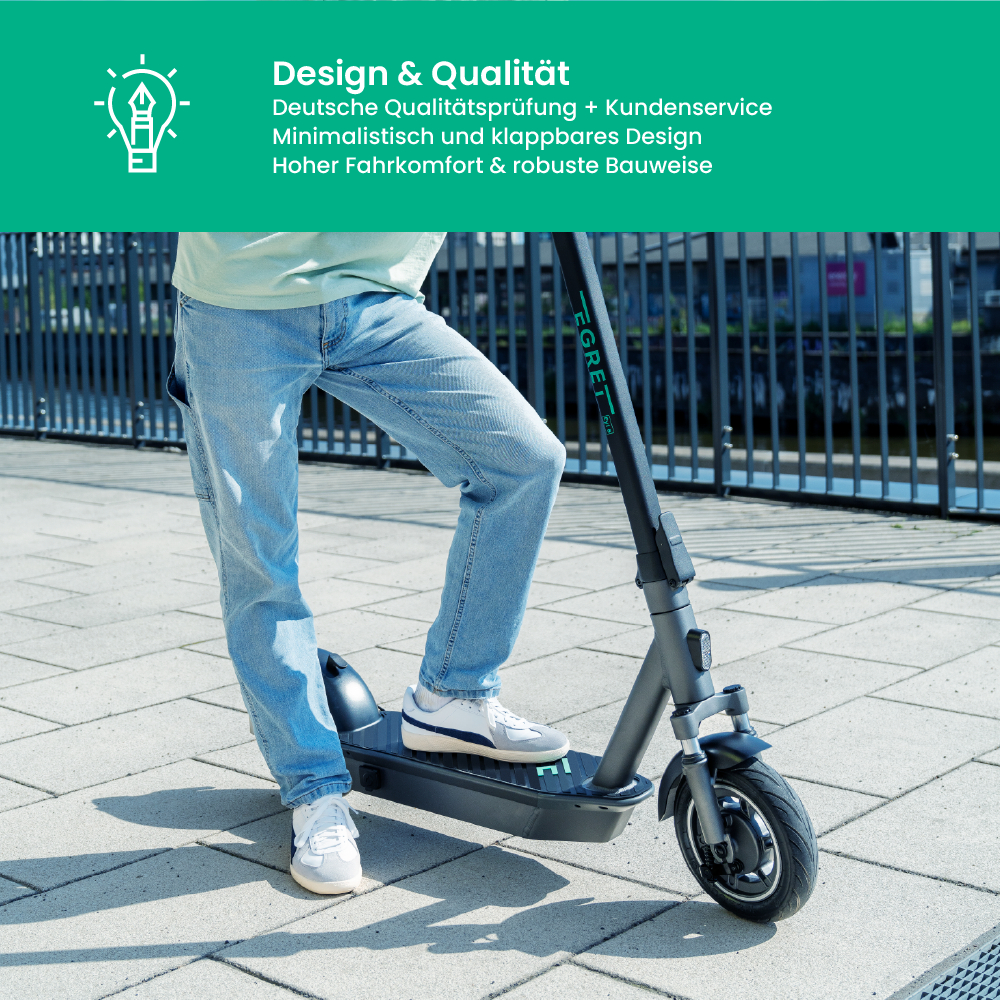 Eine Person steht auf einem grauen Elektroroller. Sie trägt Jeans und weiße Turnschuhe. Der Hintergrund zeigt ein Gebäude und einen Zaun.