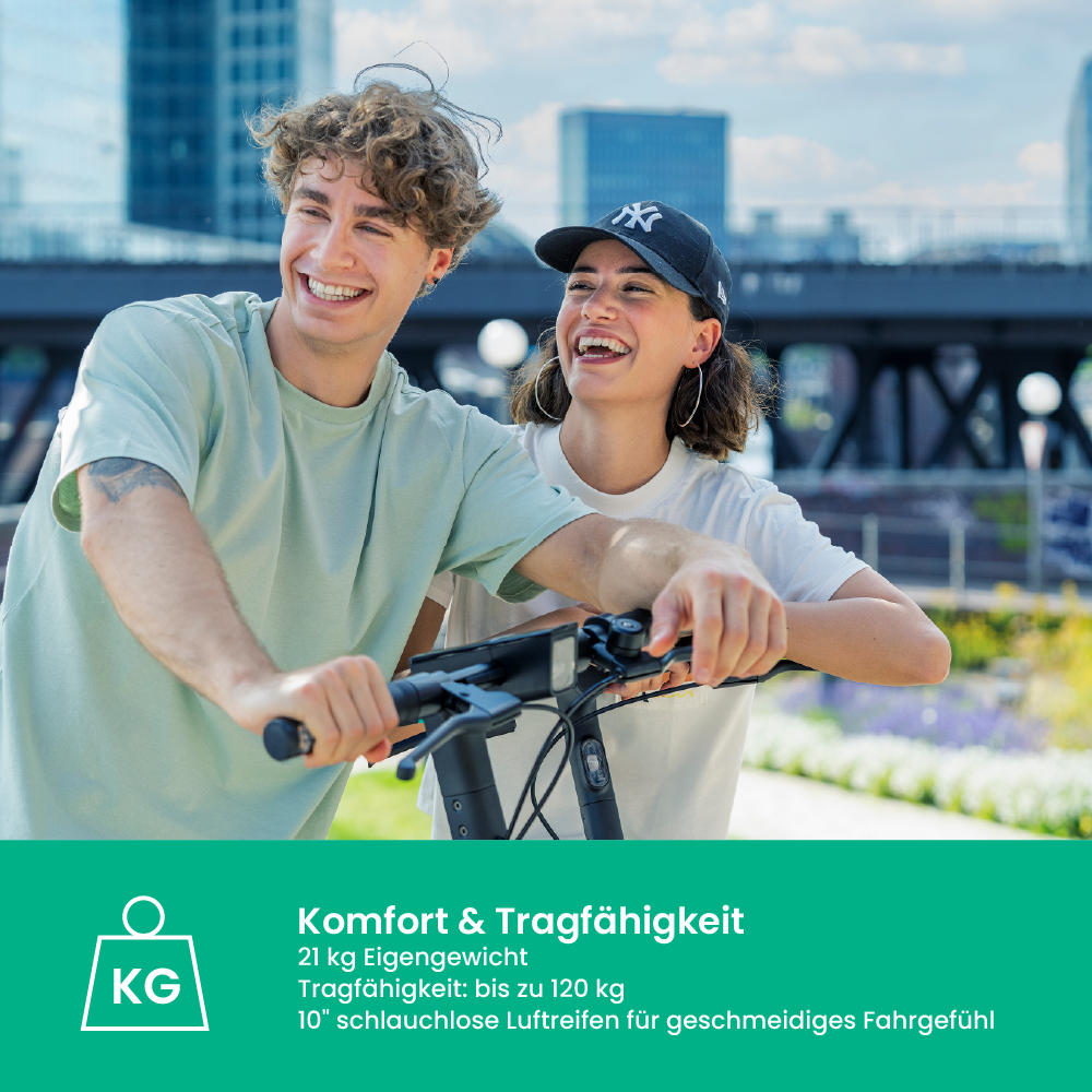 Zwei lächelnde Personen stehen mit einem E-Scooter. Der Mann trägt ein grünes Hemd, die Frau ein weißes und eine schwarze Mütze. Gebäude im Hintergrund.