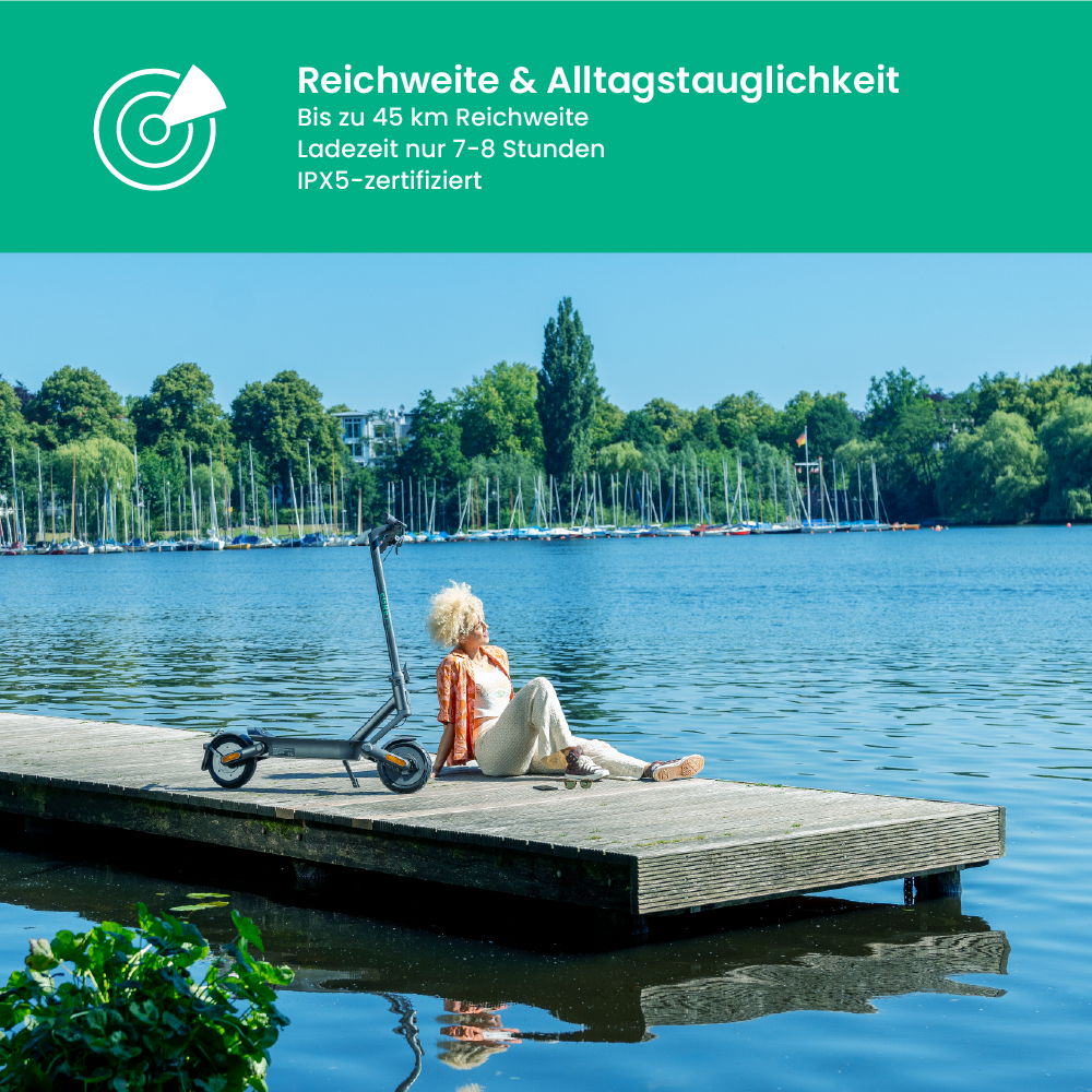 Eine Person sitzt neben einem Roller auf einem Steg. Im Hintergrund ein See mit Booten und Bäumen unter blauem Himmel.