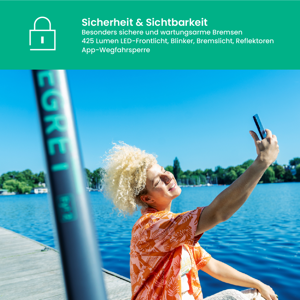 Eine Frau macht ein Selfie in der Nähe eines Sees. Sie hält ein Telefon hoch und lächelt. Der Hintergrund zeigt Wasser, Bäume und blauen Himmel.