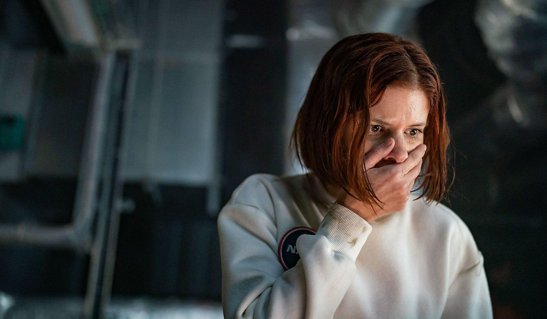 Eine Frau in einem weißen NASA-Sweatshirt bedeckt ihren Mund. Sie hat rotbraunes Haar und wirkt besorgt, mit einem dunklen, verschwommenen Hintergrund.