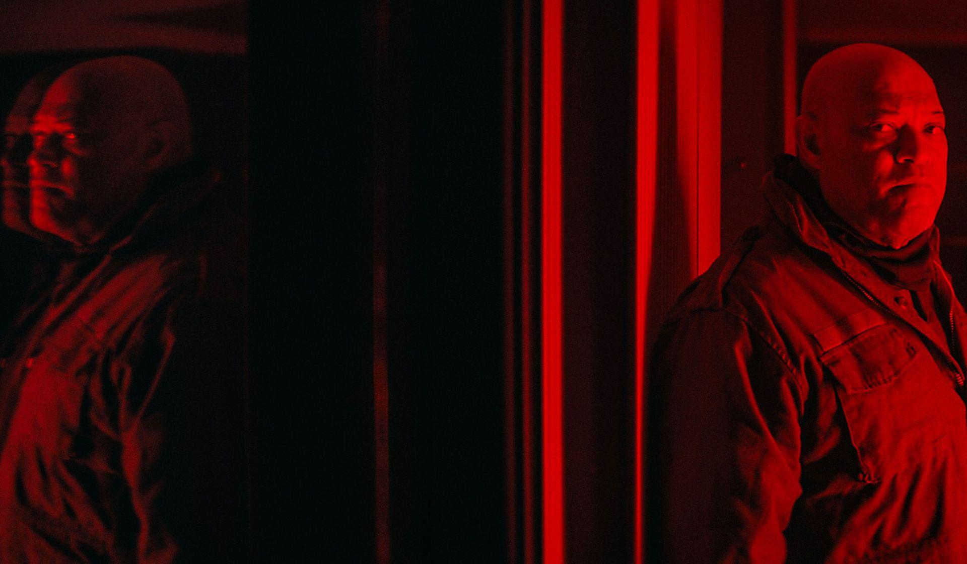 Ein Mann in roter Jacke steht in einem rot beleuchteten Raum. Ein Spiegelbild. Der Hintergrund ist dunkel.