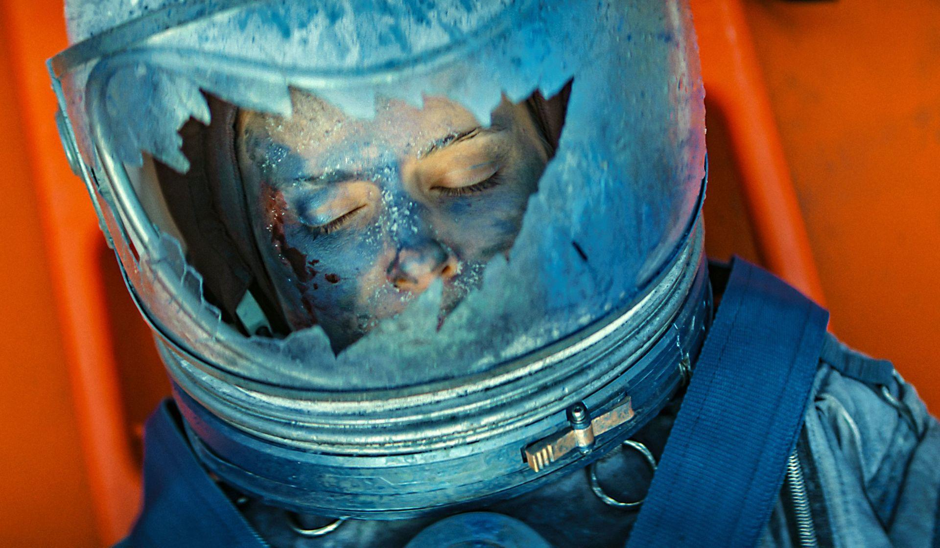 Eine Person in einem beschädigten Astronautenhelm. Das Gesicht ist mit geschlossenen Augen sichtbar. Blaue und weiße Farbe befindet sich im Gesicht und auf dem Helm.