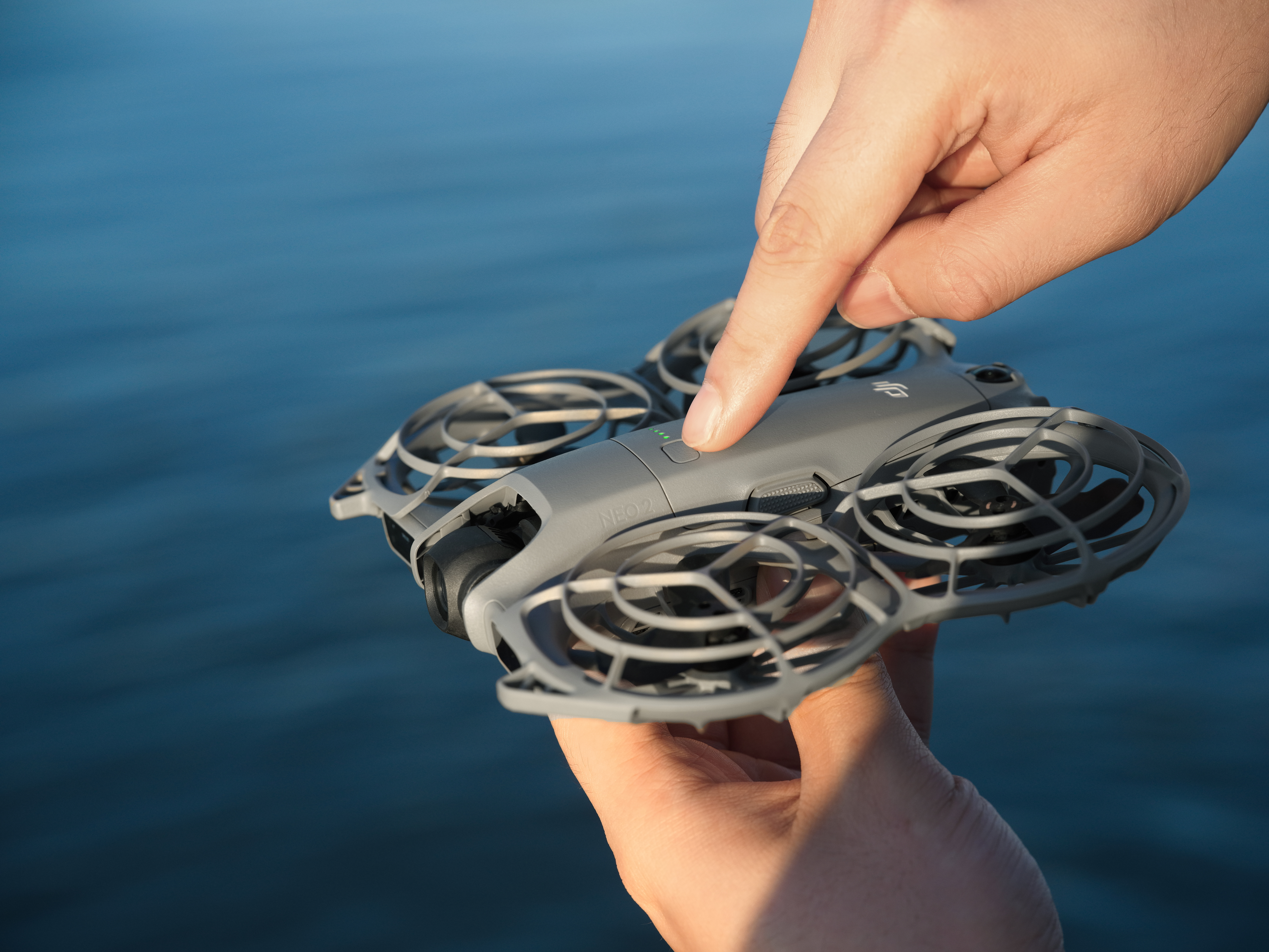 Die Hand einer Person drückt auf den Einschaltknopf einer Drohne. Die Drohne ist grau, der Hintergrund blaues Wasser.