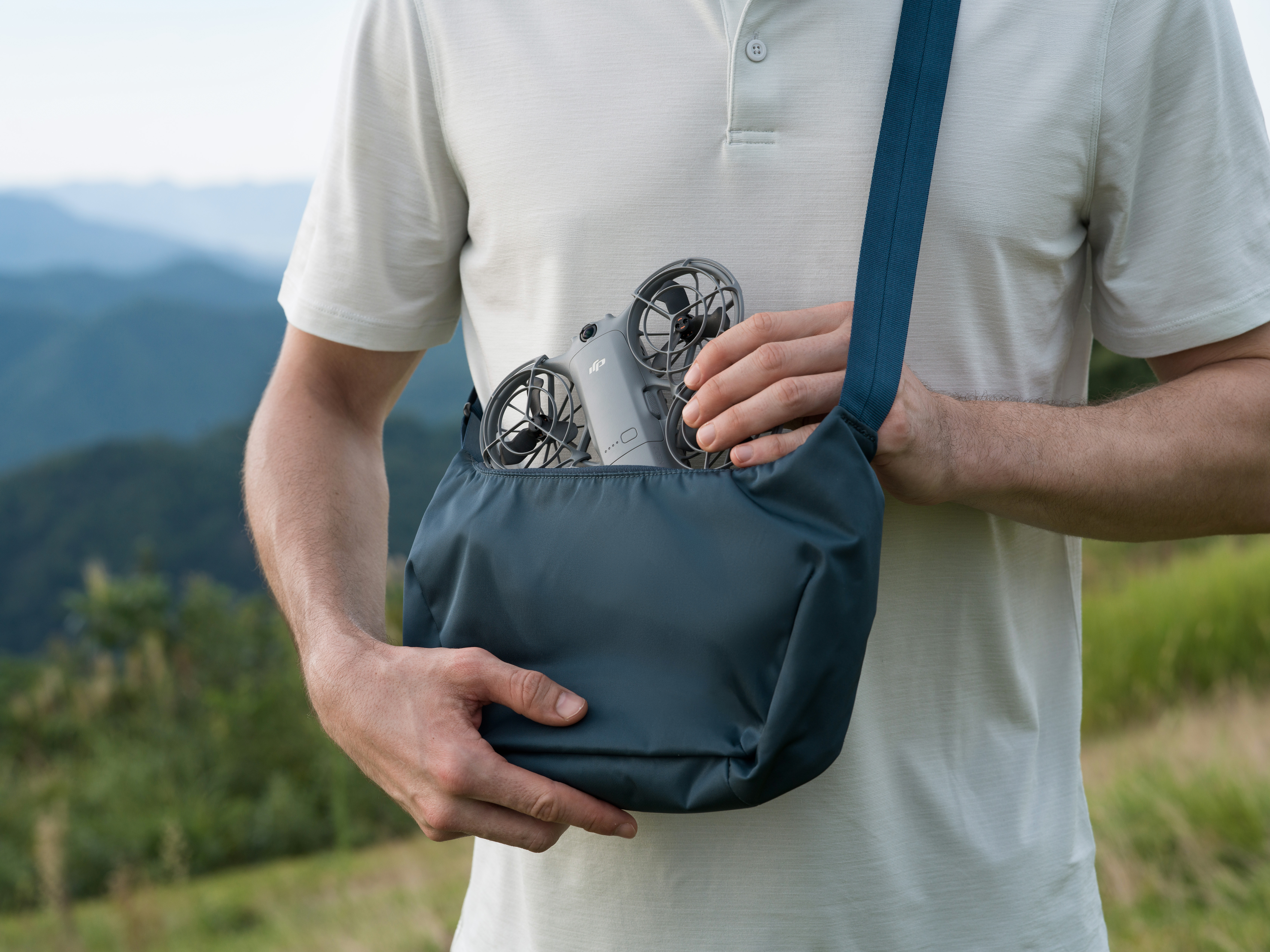 Eine Person hält eine Drohne in einer blauen Tasche. Im Hintergrund sind Berge und Gras zu sehen.