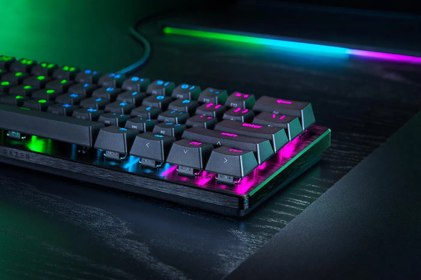 Egy fekete Razer billentyűzet világító gombokkal. Zöld, kék és rózsaszín fények láthatók. Sötét fa felületen fekszik.