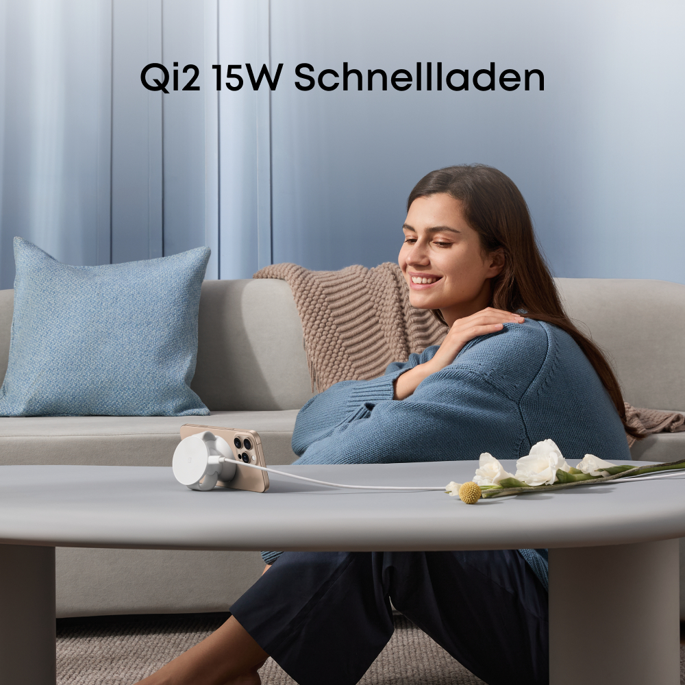 Eine Frau lächelt, während sie ihr Telefon auflädt. Ein weißes Ladegerät liegt auf dem Tisch, Blumen und ein Kissen sind in der Nähe.