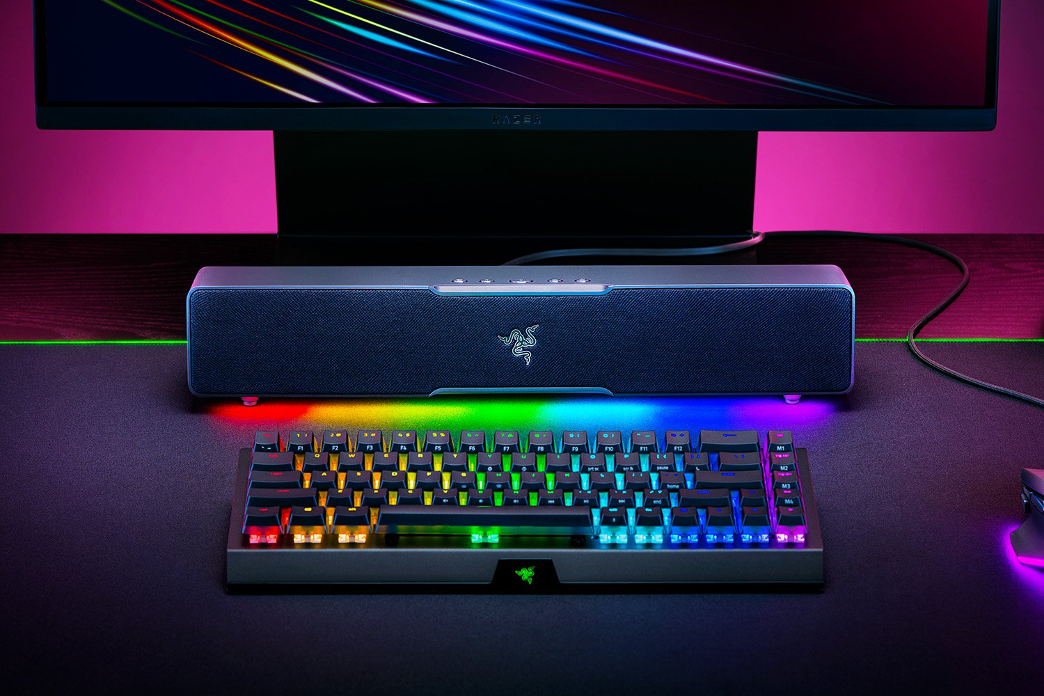 Egy gamer beállítás billentyűzettel, hangszóróval és monitorral. A billentyűzet többszínűen világít. A monitor színes csíkokat jelenít meg.