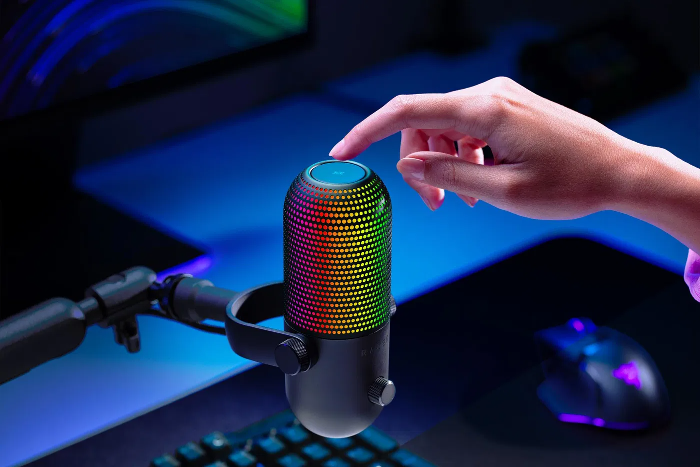 Egy kéz megnyom egy gombot egy RGB világítású mikrofonon. Az egér és a billentyűzet a homályos háttérben.