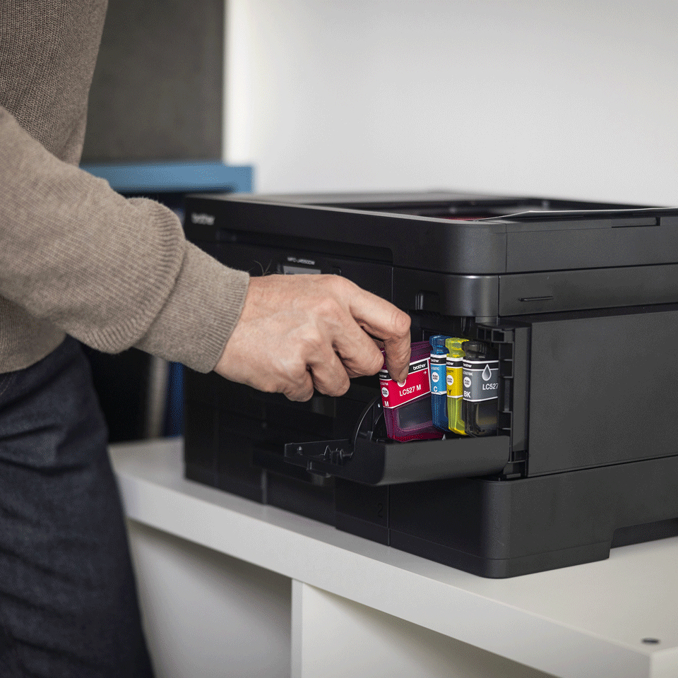 Eine Person setzt eine magentafarbene Tintenpatrone in einen schwarzen Brother-Drucker ein. Weitere Patronen sind sichtbar. Der Drucker steht auf einem weißen Tisch.