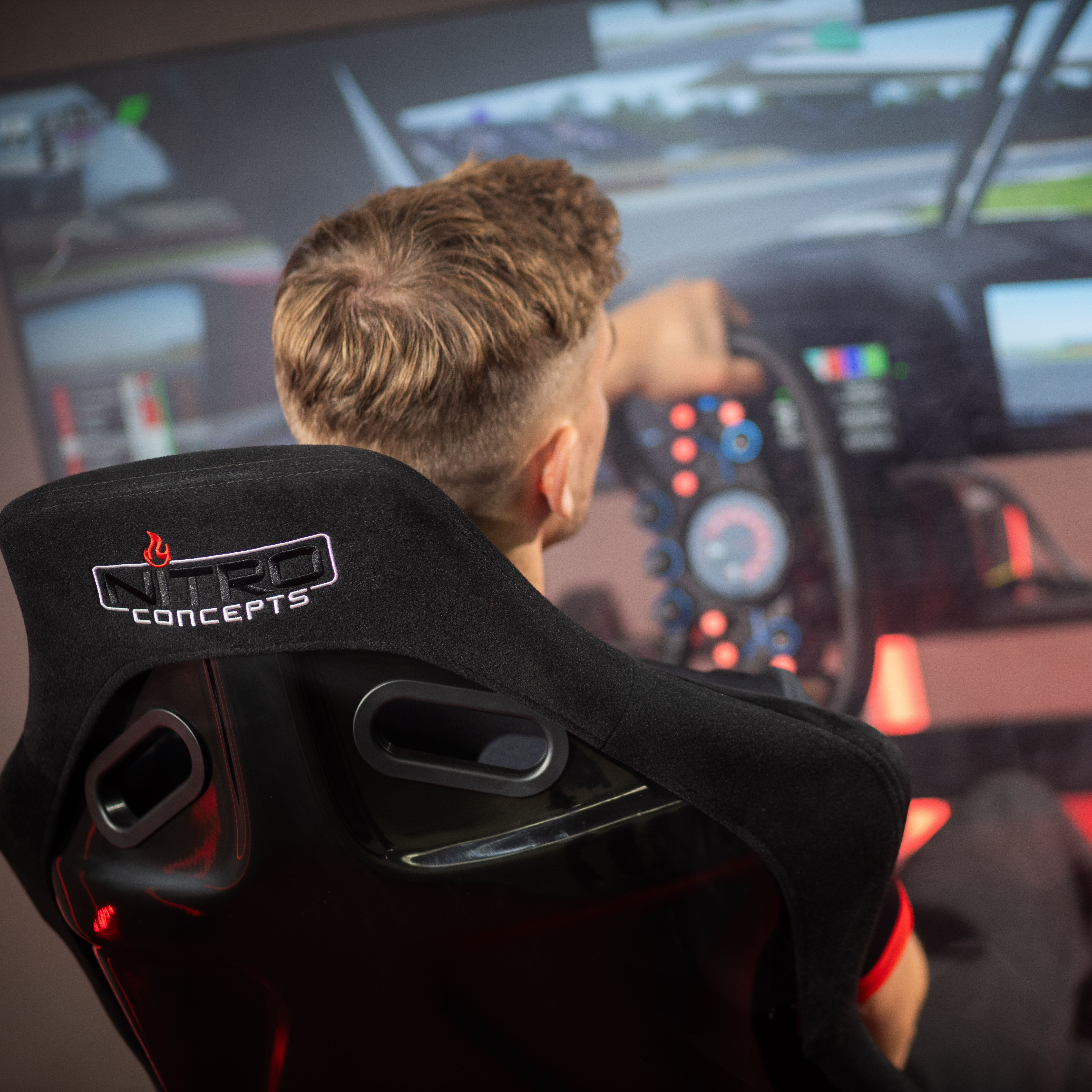 Eine Person in einem Rennsimulator. Die Ansicht ist von hinten, mit schwarzem Sitz und Rennspiel-Bildschirm.