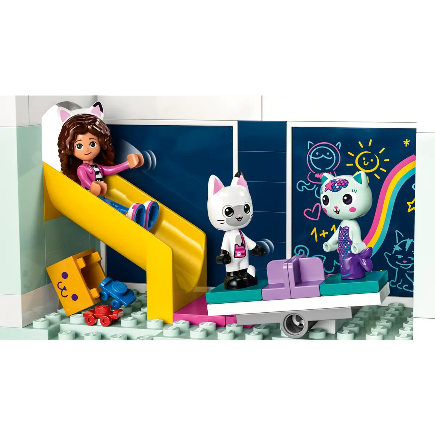 Een Lego-set met een meisje dat van een gele glijbaan glijdt, twee kattenfiguren en een schoolbord met tekeningen.