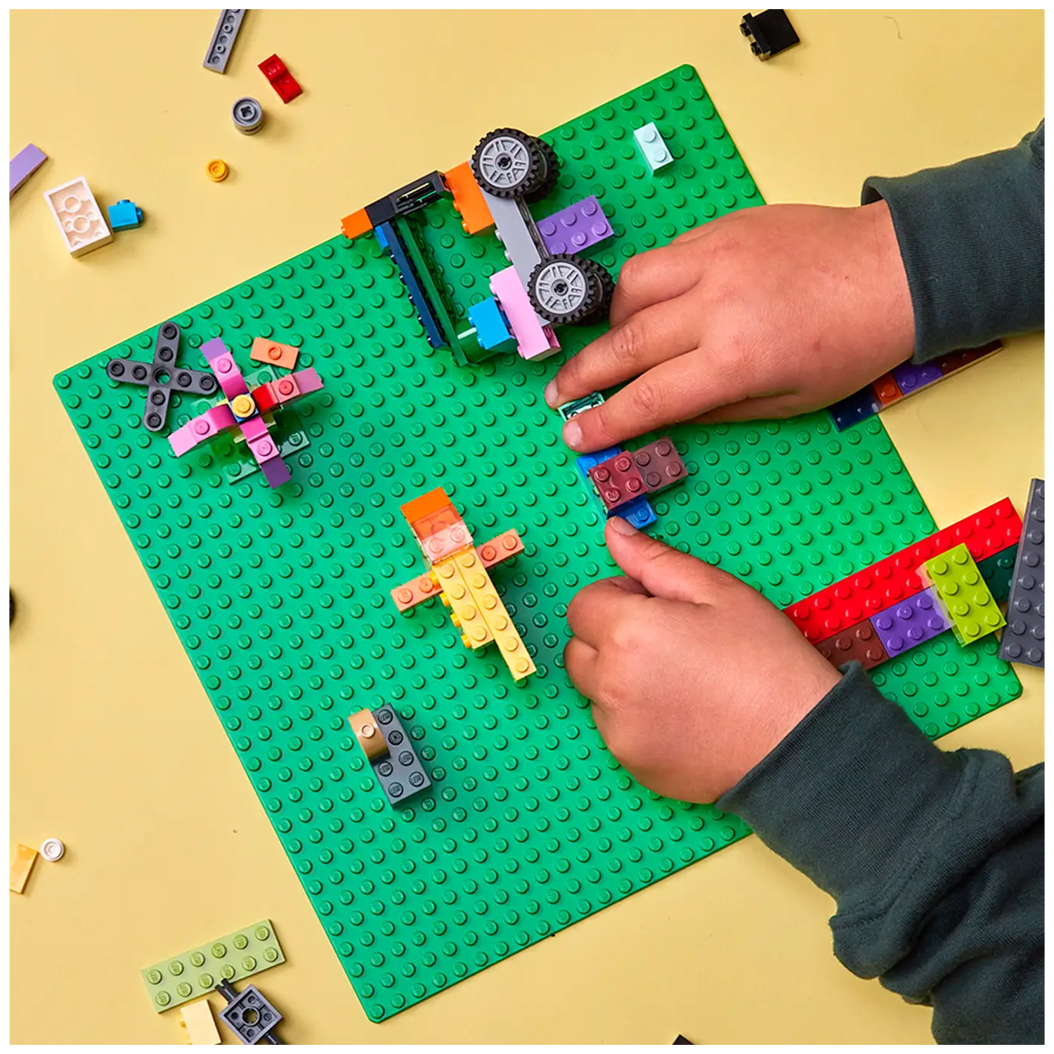 Hands building with Lego bricks on a green baseplate. Various Lego creations, including a car and a person. Yellow background.