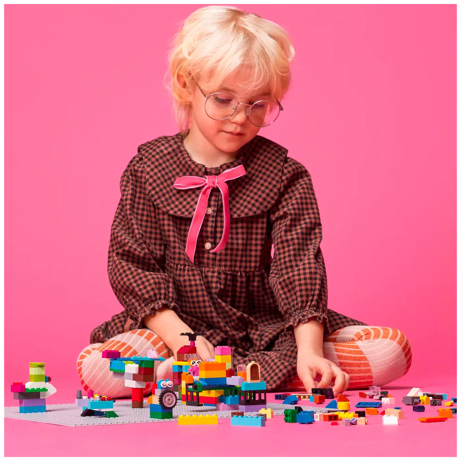 A child with glasses and blonde hair sits on the floor, playing with colorful building blocks against a pink background.