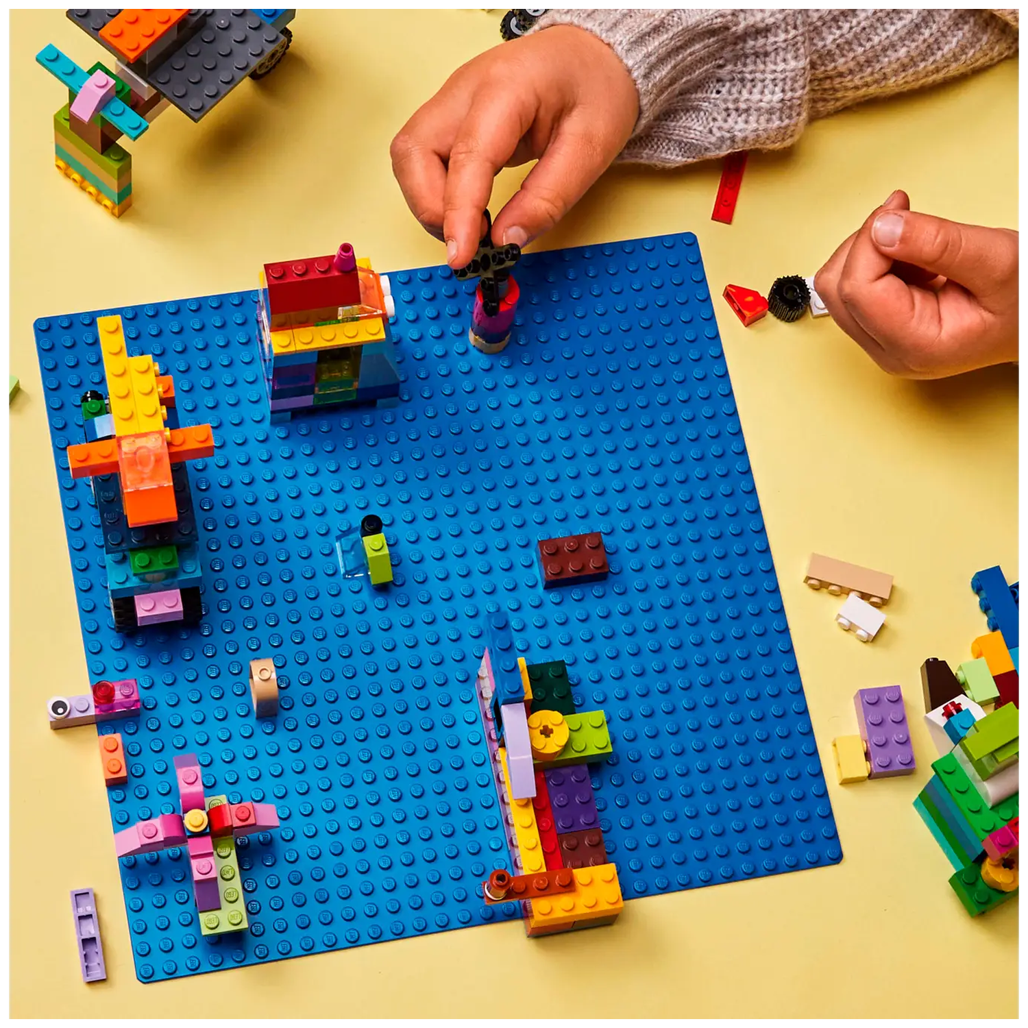 Hands building with colorful LEGO bricks on a blue baseplate. Various structures are partially built. Yellow background.