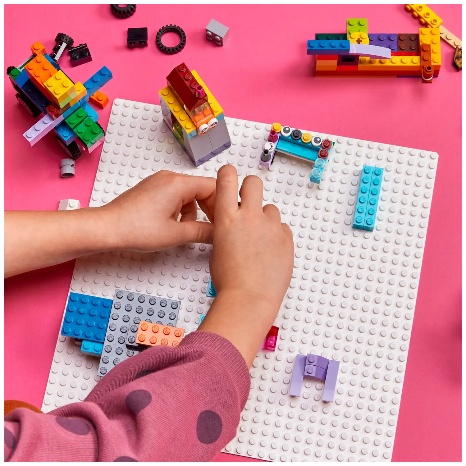 Handen van een kind bouwen met kleurrijke Lego-stenen op een witte basisplaat tegen een roze achtergrond.