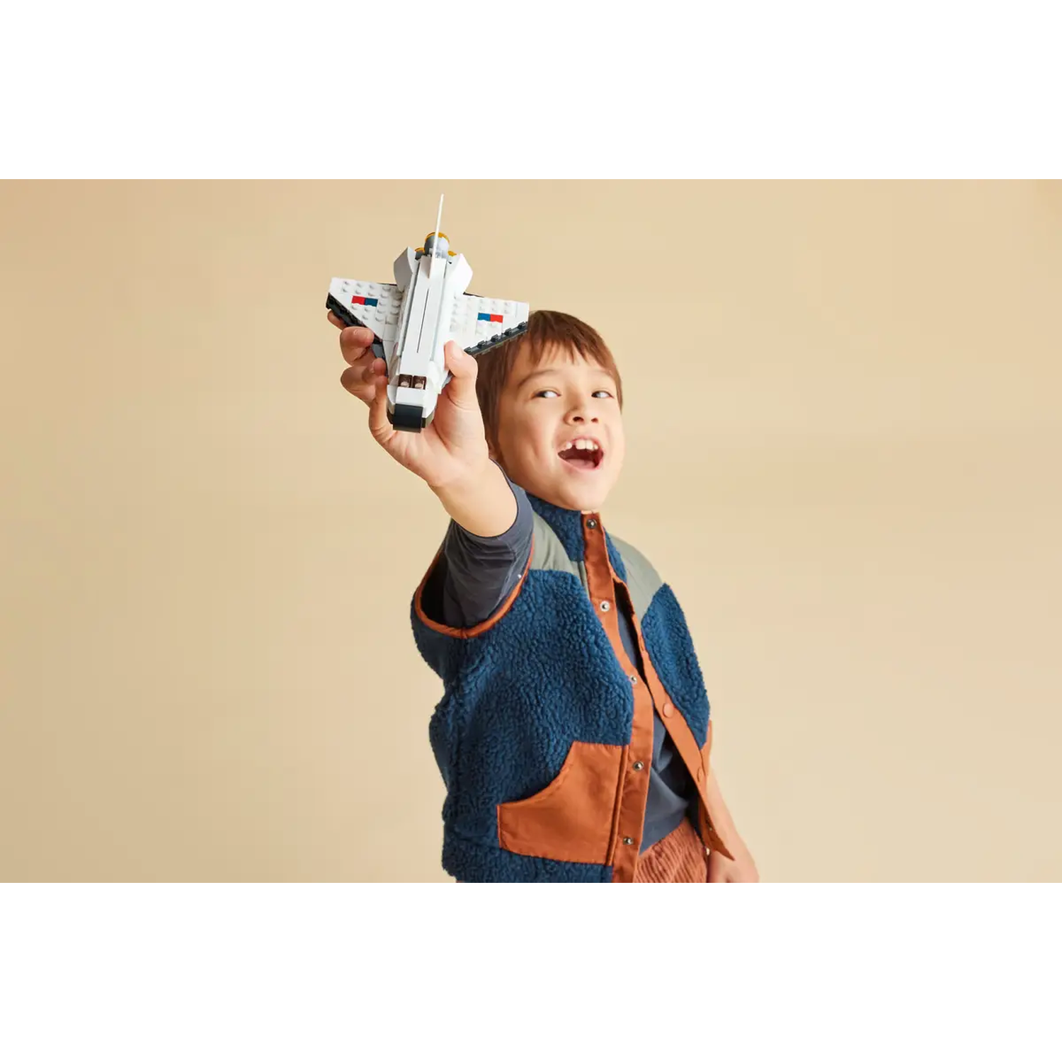 A boy holds up a toy space shuttle. He wears a blue and brown vest, looking excited. Beige background.