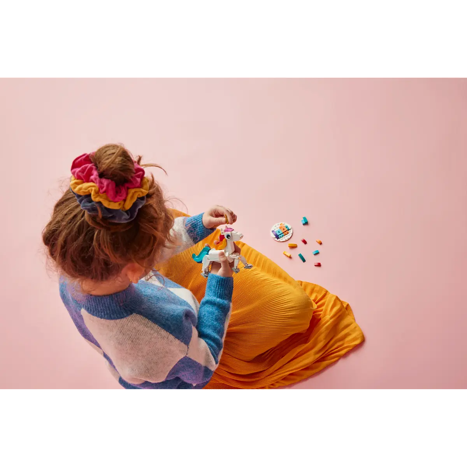 A child in a blue sweater and yellow skirt holds a toy unicorn. Colorful scrunchies in hair. Pink background with toys.