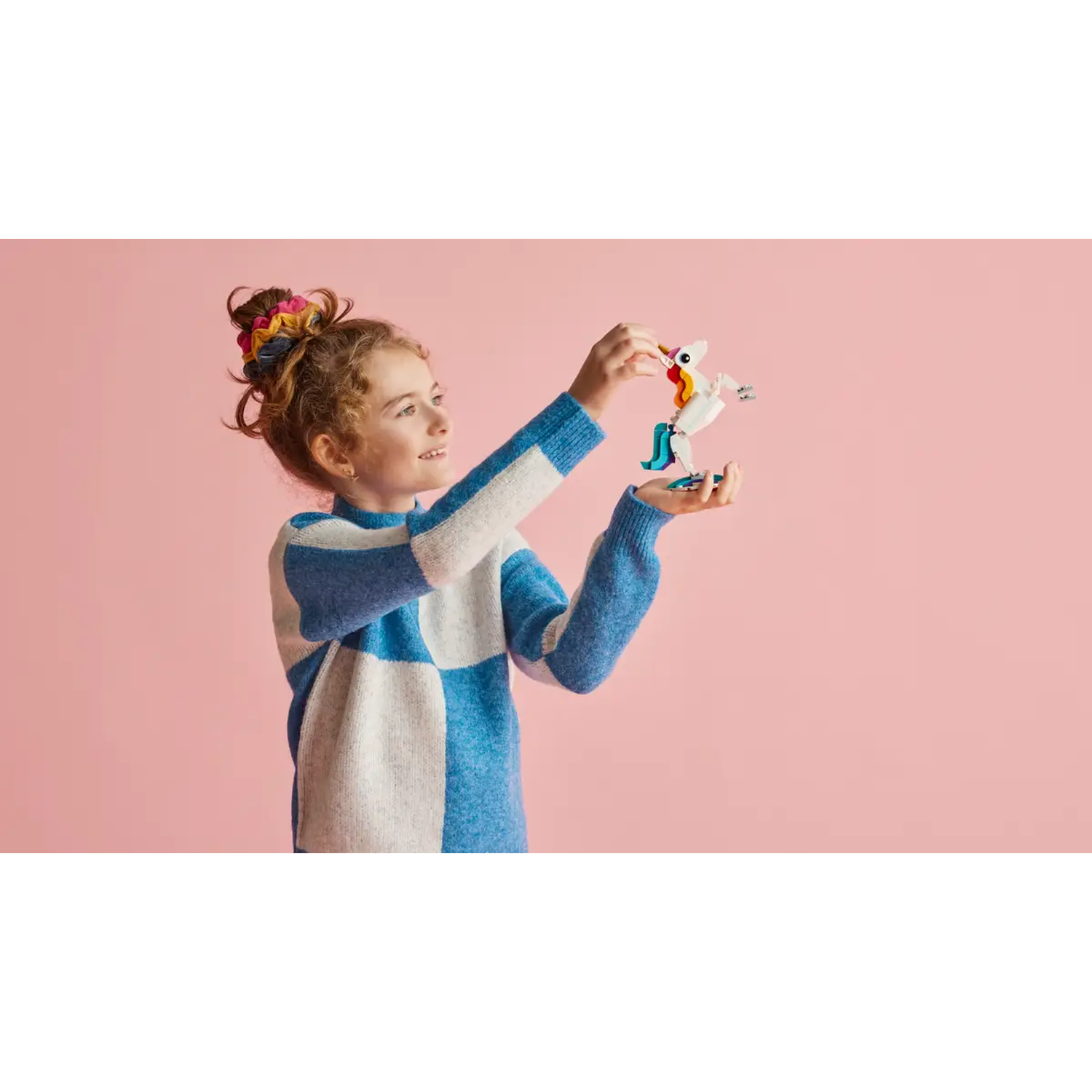 A girl in a blue and white sweater holds a toy unicorn against a pink background. She looks up smiling.