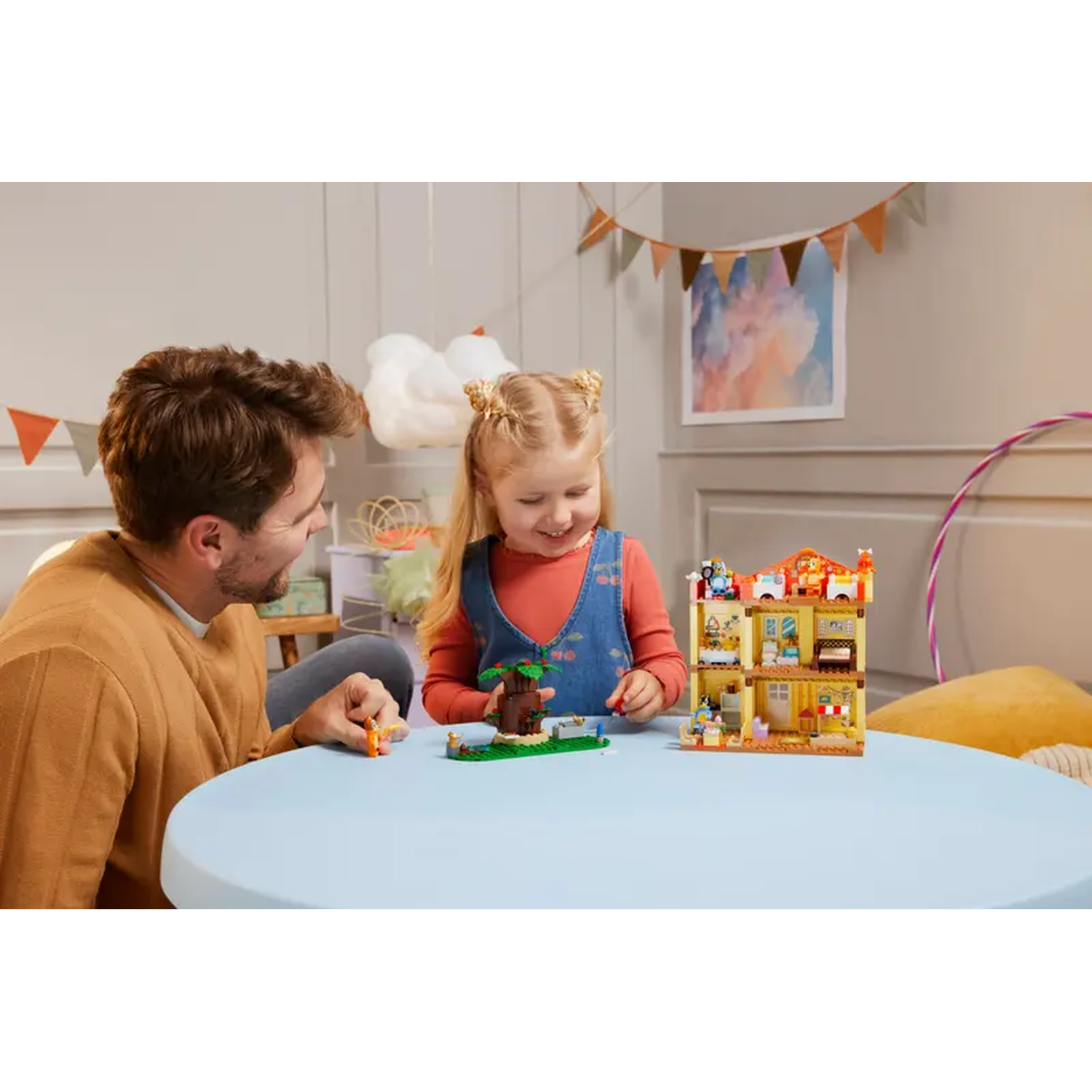 Een man en een meisje spelen met een speelgoedhuis. Het meisje lacht. Er staat een boom en wat speelgoed op een blauwe tafel.