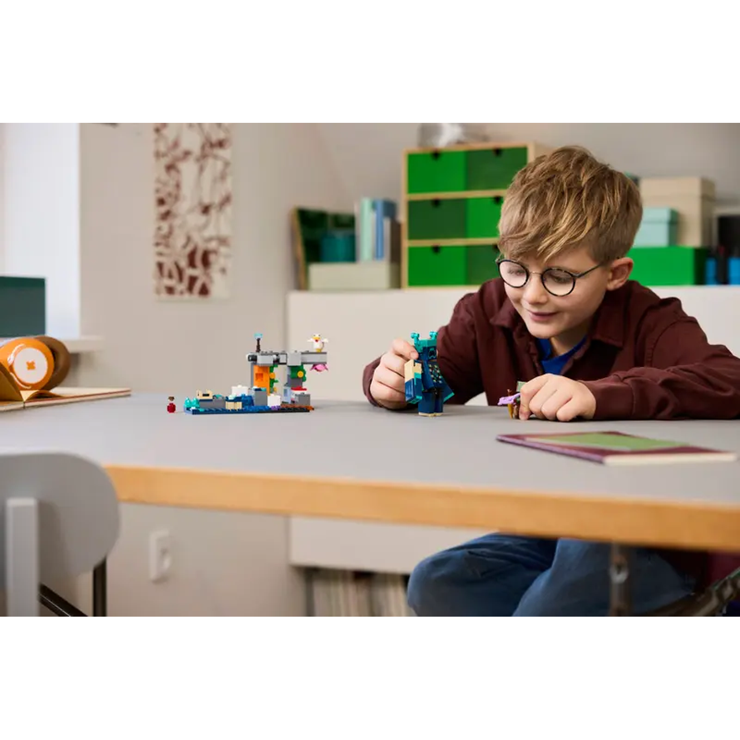 Een jongen met een bril speelt met LEGO's. Kleurrijke LEGO-structuren en figuren staan op een grijze tafel. Achtergrond: een muur en opbergruimte.