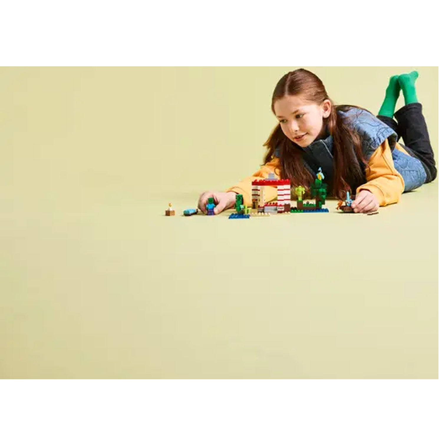A girl is laying on the floor, looking at a Lego set. The set has a building, trees, and other objects.