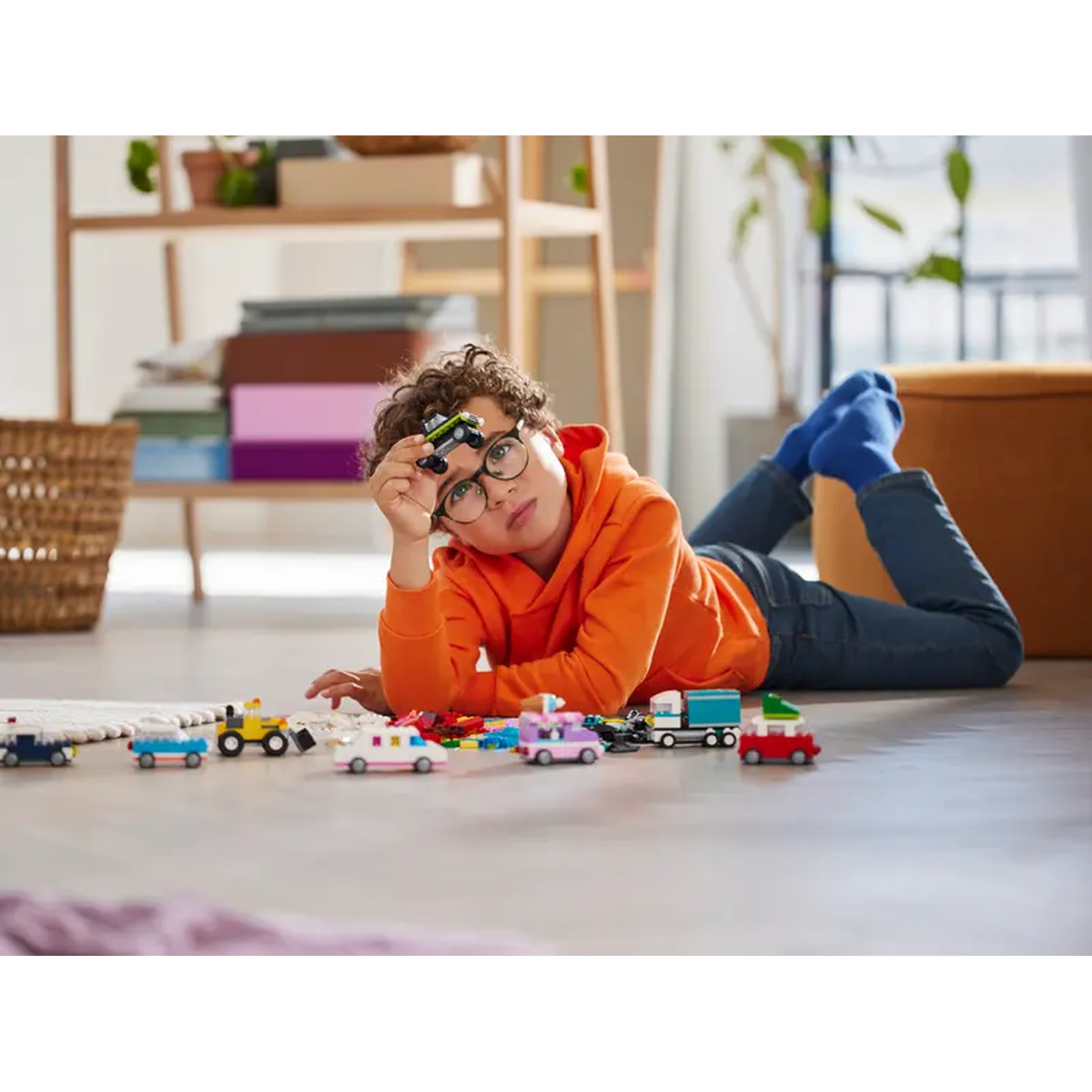 A boy in orange hoodie lies on floor, playing with toy cars. Cars of different colors are scattered around. A shelf and a basket are in the background.