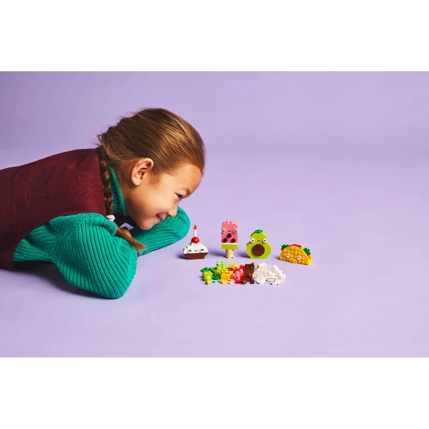 A girl looks at Lego food: cake, watermelon popsicle, avocado, and taco. Colorful bricks are scattered on a purple surface.