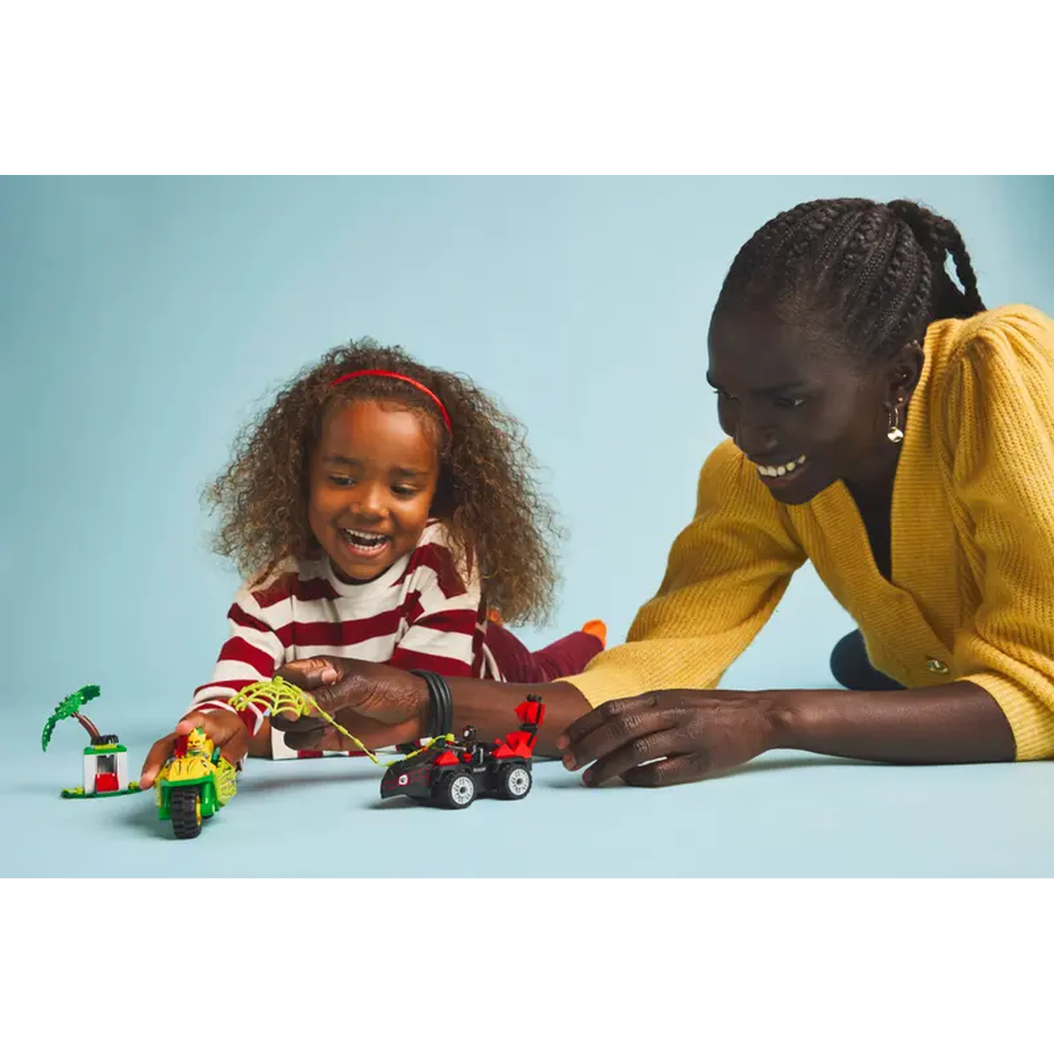 A girl and woman play with toy vehicles on a blue surface. The girl wears a red headband and striped shirt. The woman wears a yellow sweater.