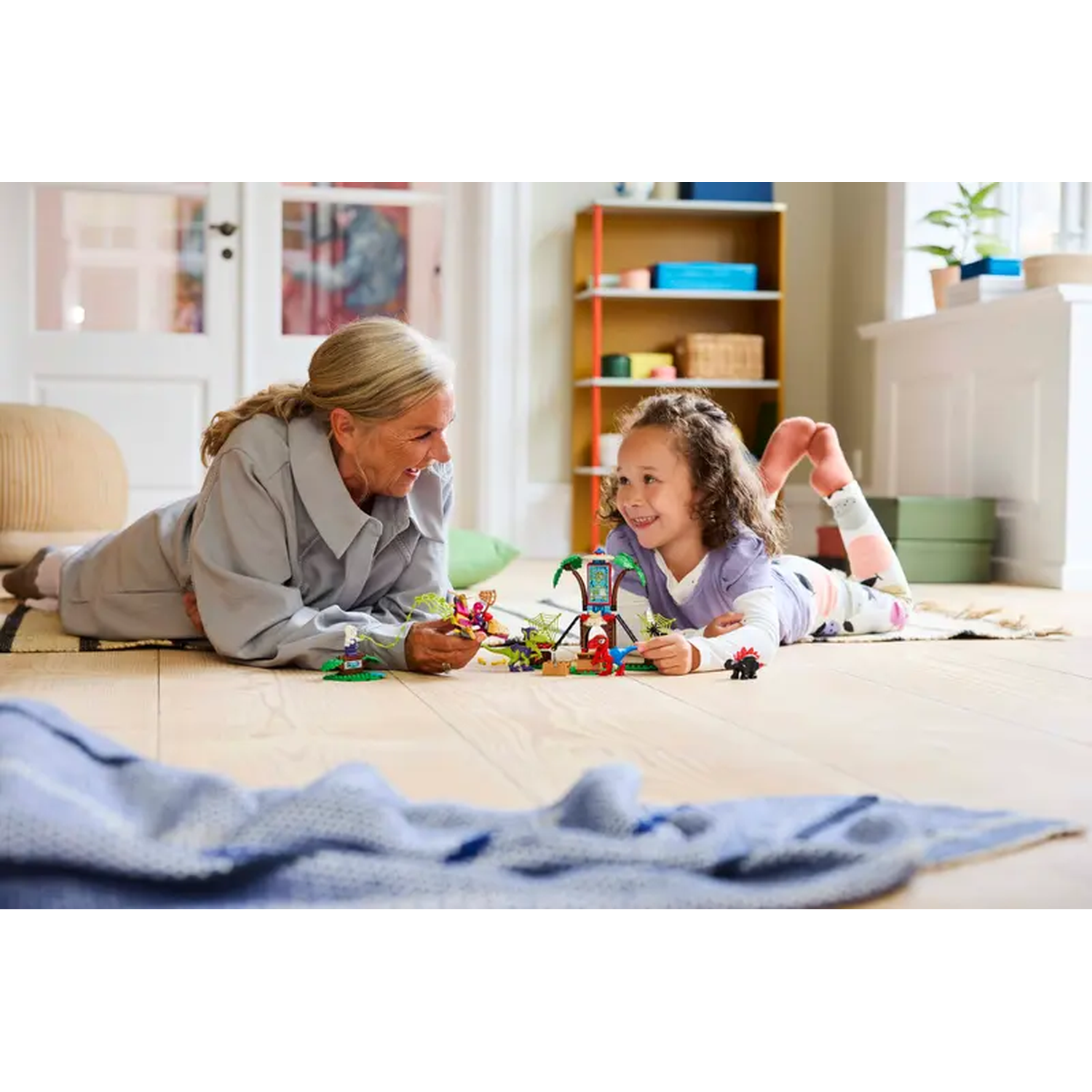 A woman and a girl are playing with toys on the floor. A blue blanket is in the foreground. A shelf is in the background.