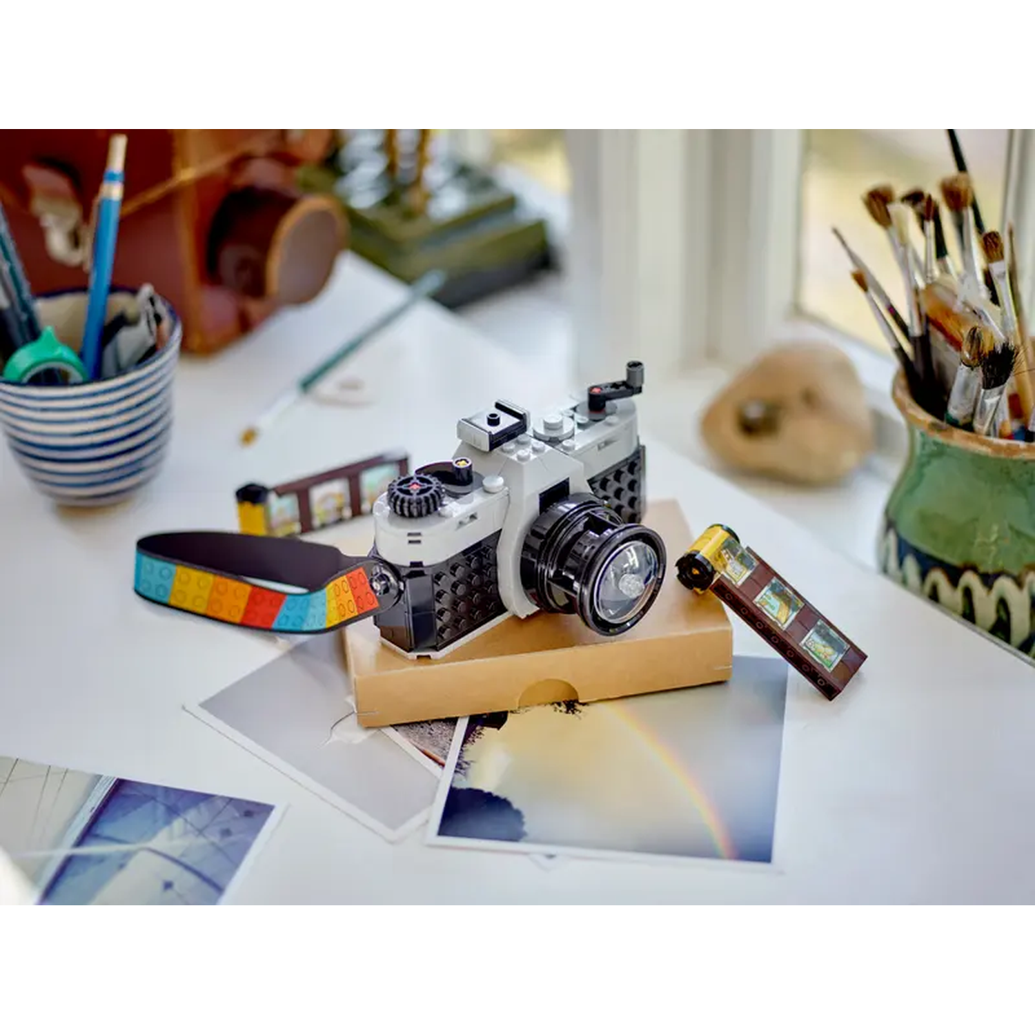 A Lego camera set on a white table. It has a rainbow film strip, pictures, and pencils in a cup. Paint brushes are in a vase.