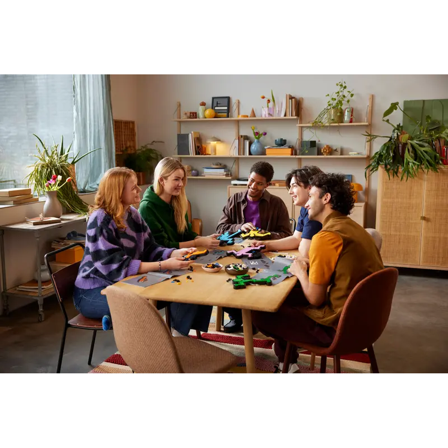 Five people sit around a table assembling toys. The room is bright, with plants, a bookcase, and a window with a curtain.