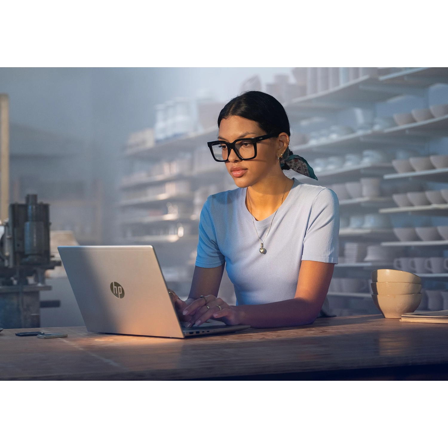 Une femme avec des lunettes tape sur un ordinateur portable HP. Elle porte une chemise bleue. Des étagères avec de la poterie blanche en arrière-plan.