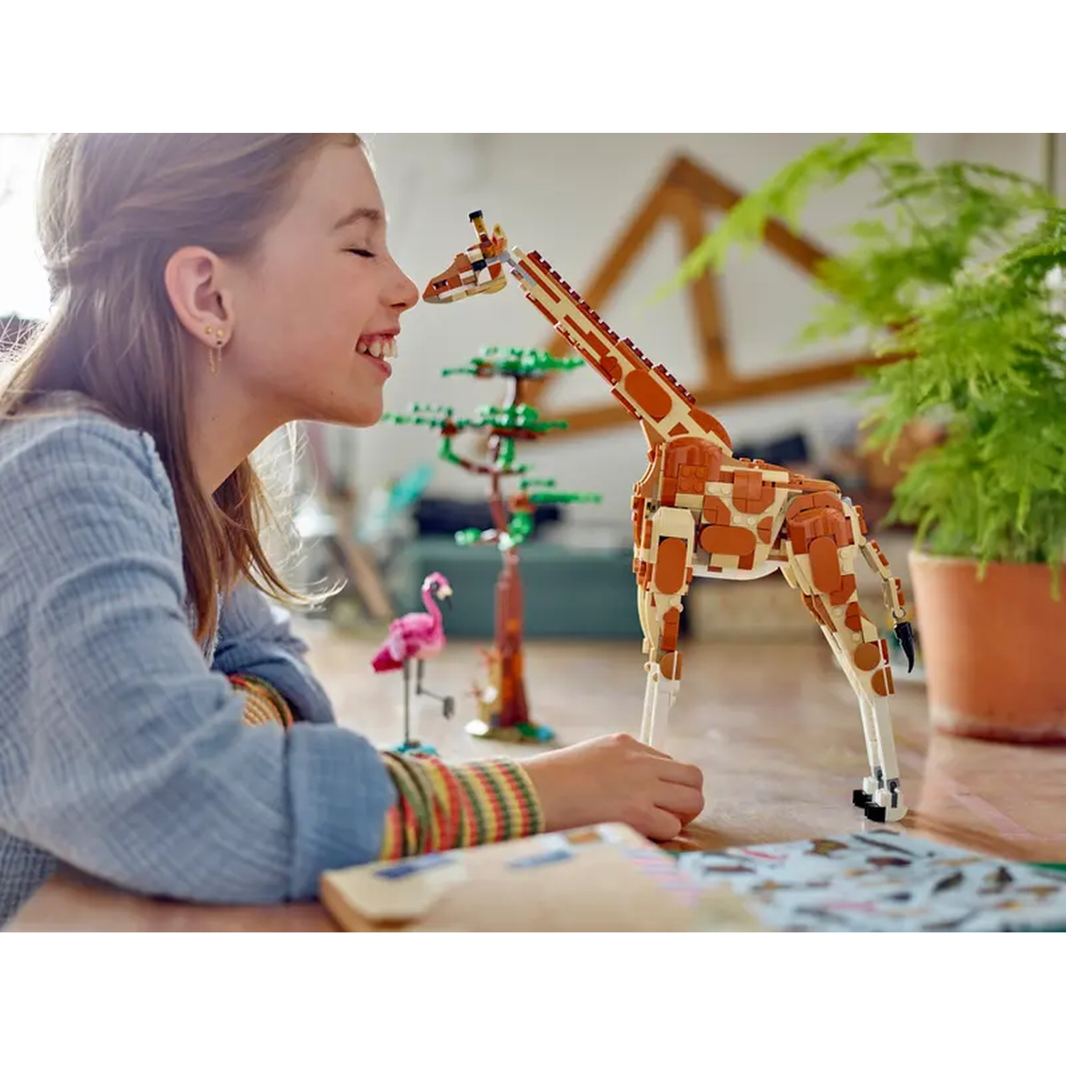 A girl smiles at a Lego giraffe. Flamingo and tree Lego models are in the background, near a fern plant.
