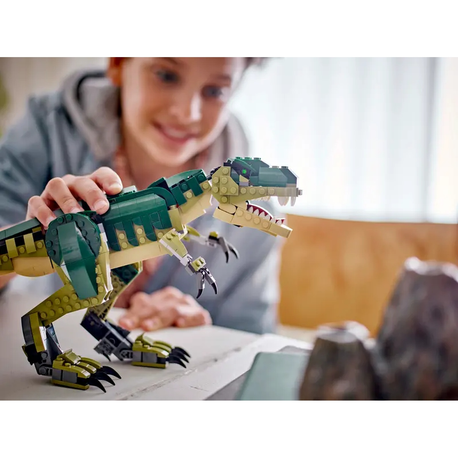 A smiling person plays with a Lego dinosaur model. The dinosaur is green and tan. The background has a blurred view.
