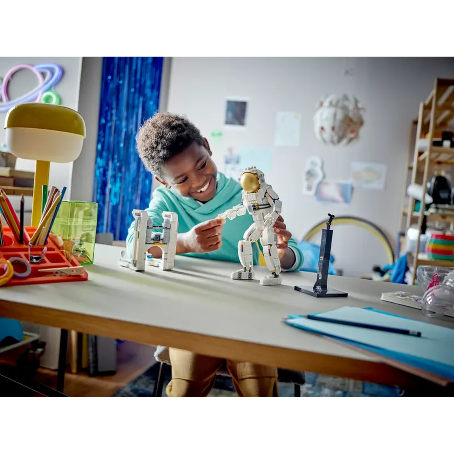 A young boy smiles, playing with a Lego astronaut. Colorful desk items and decor are in the background, including a neon sign.