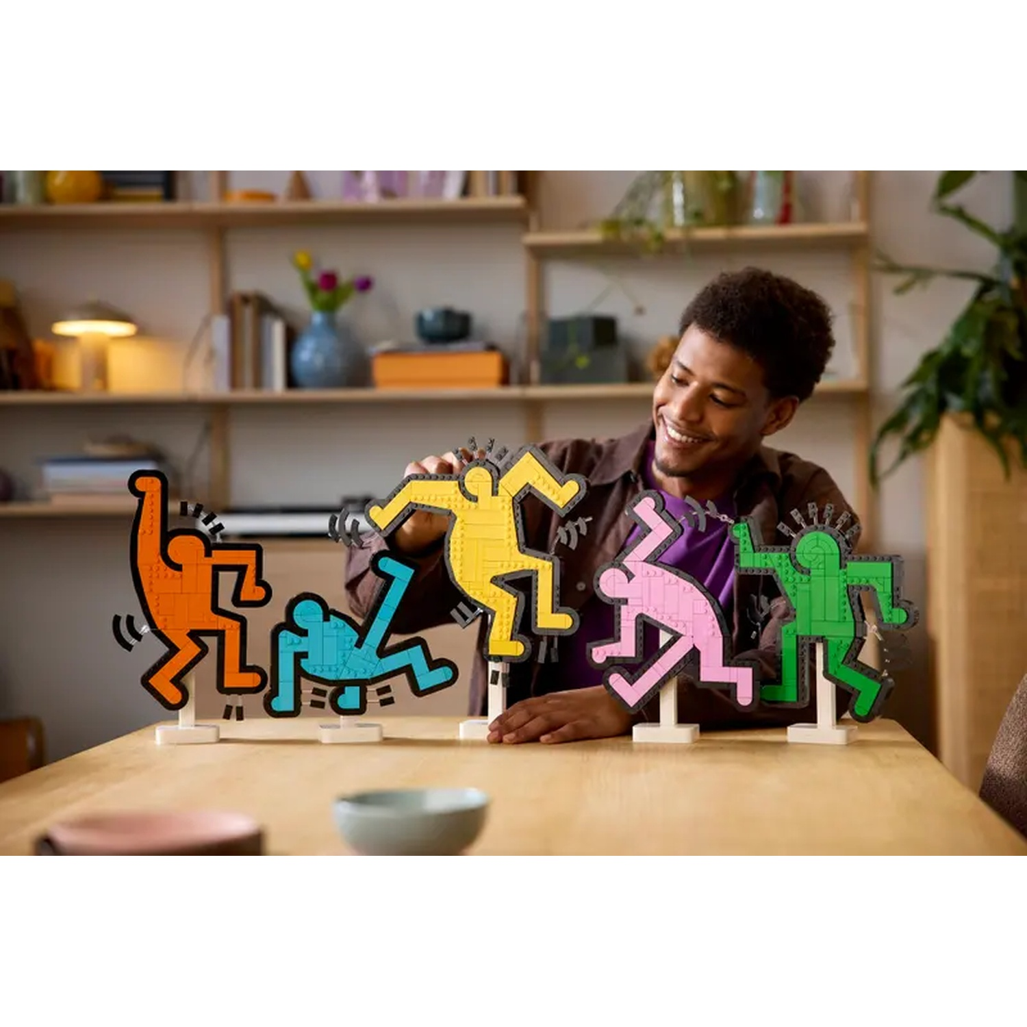 A man smiles, arranging colorful Lego figures on a table. Figures are orange, blue, yellow, pink, and green.