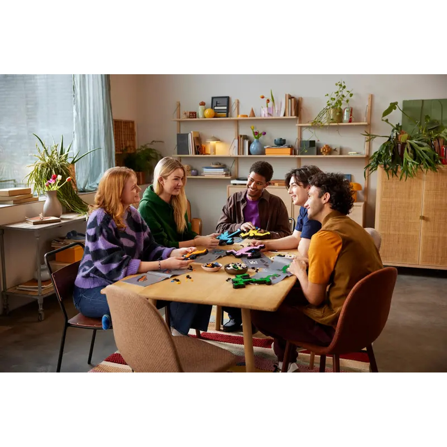 Five people sit around a table assembling toys. A variety of colored toys are on the table. The room has wooden furniture and plants.