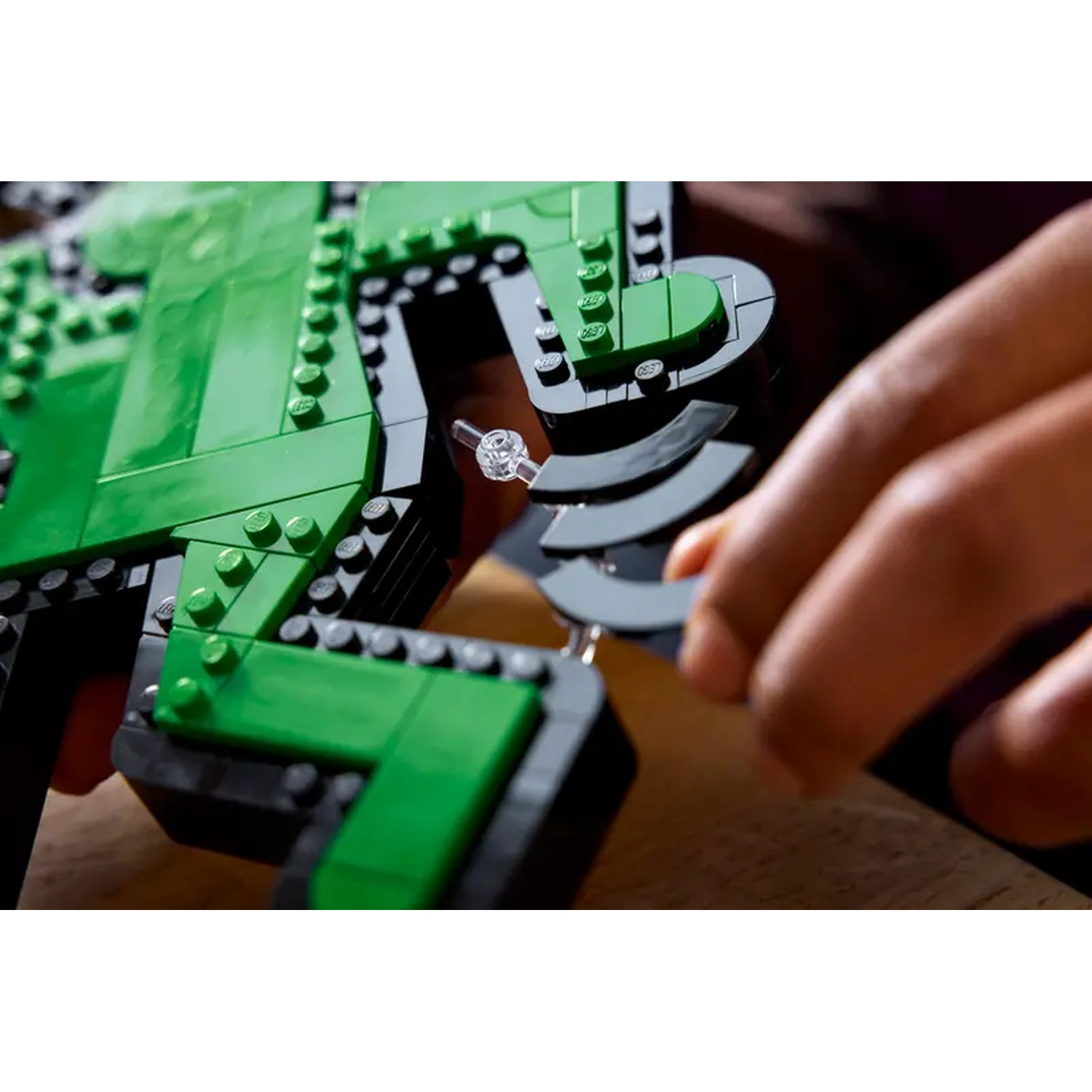Hands assembling a green and black Lego structure. Clear plastic piece is being connected. Wooden surface visible.